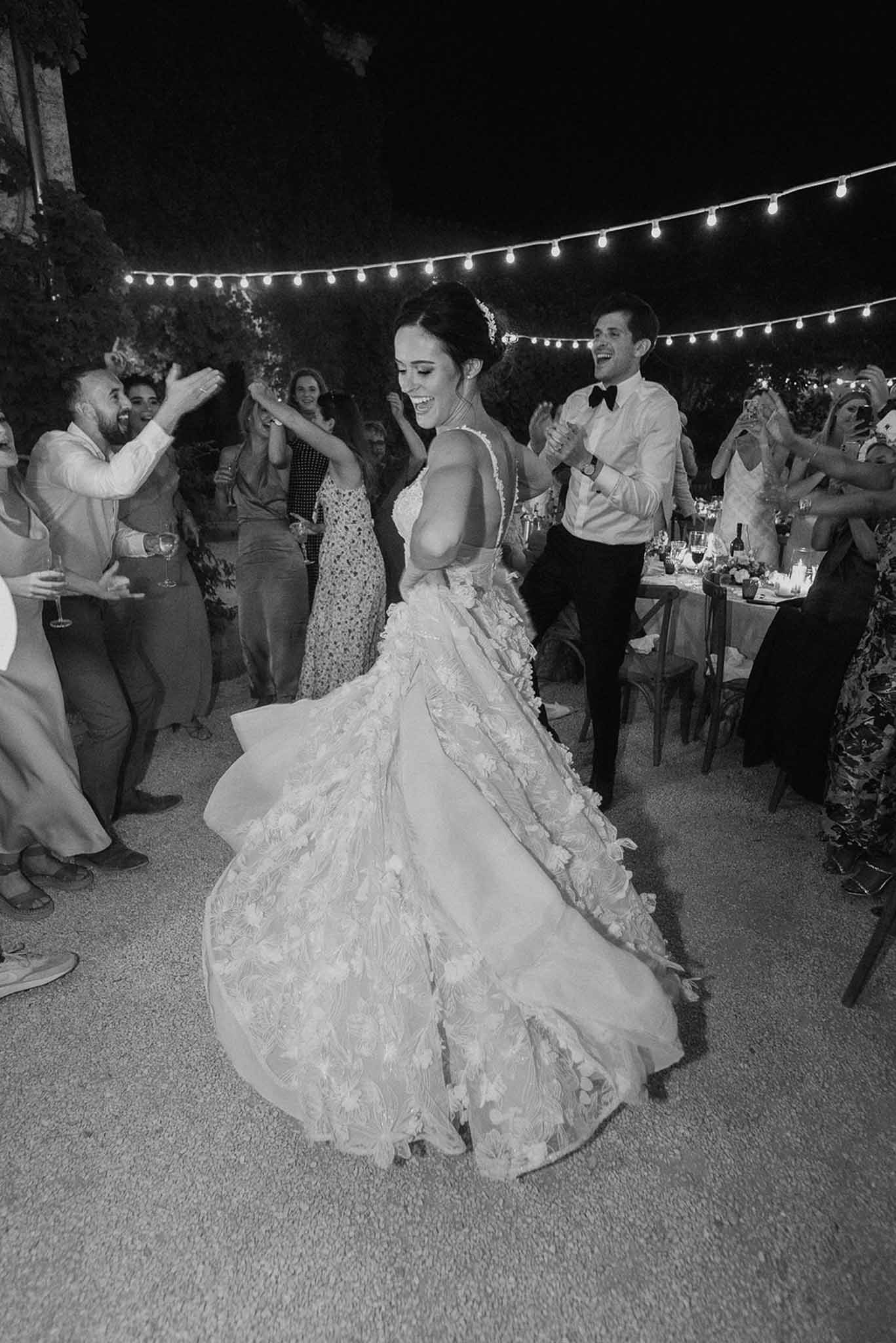 Bride spinning in floral-applique ballgown as groom claps and guests circle outdoor dance floor in B&W