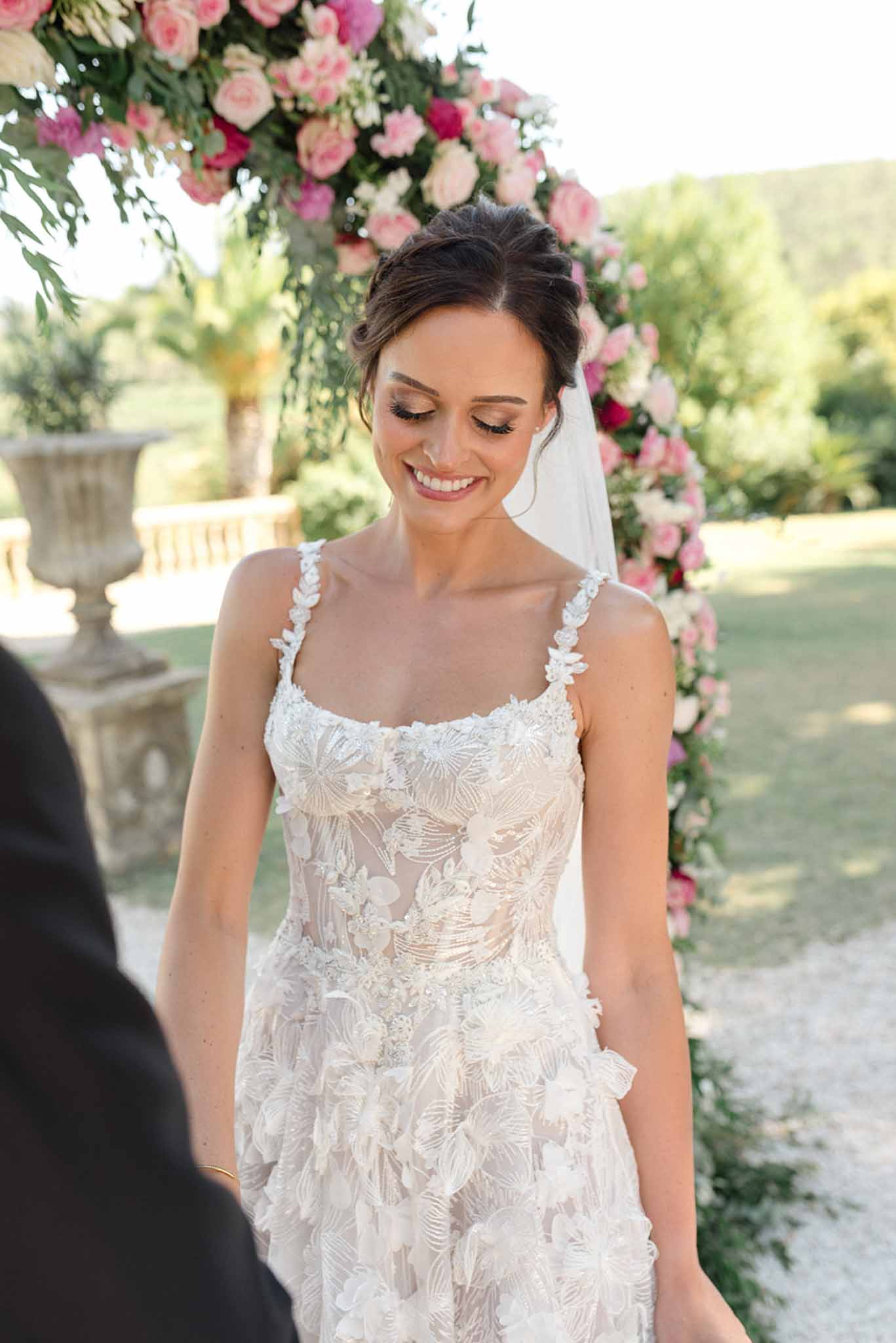 Bride smiling in sheer floral lace gown before hot pink and white rose ceremony arch in garden