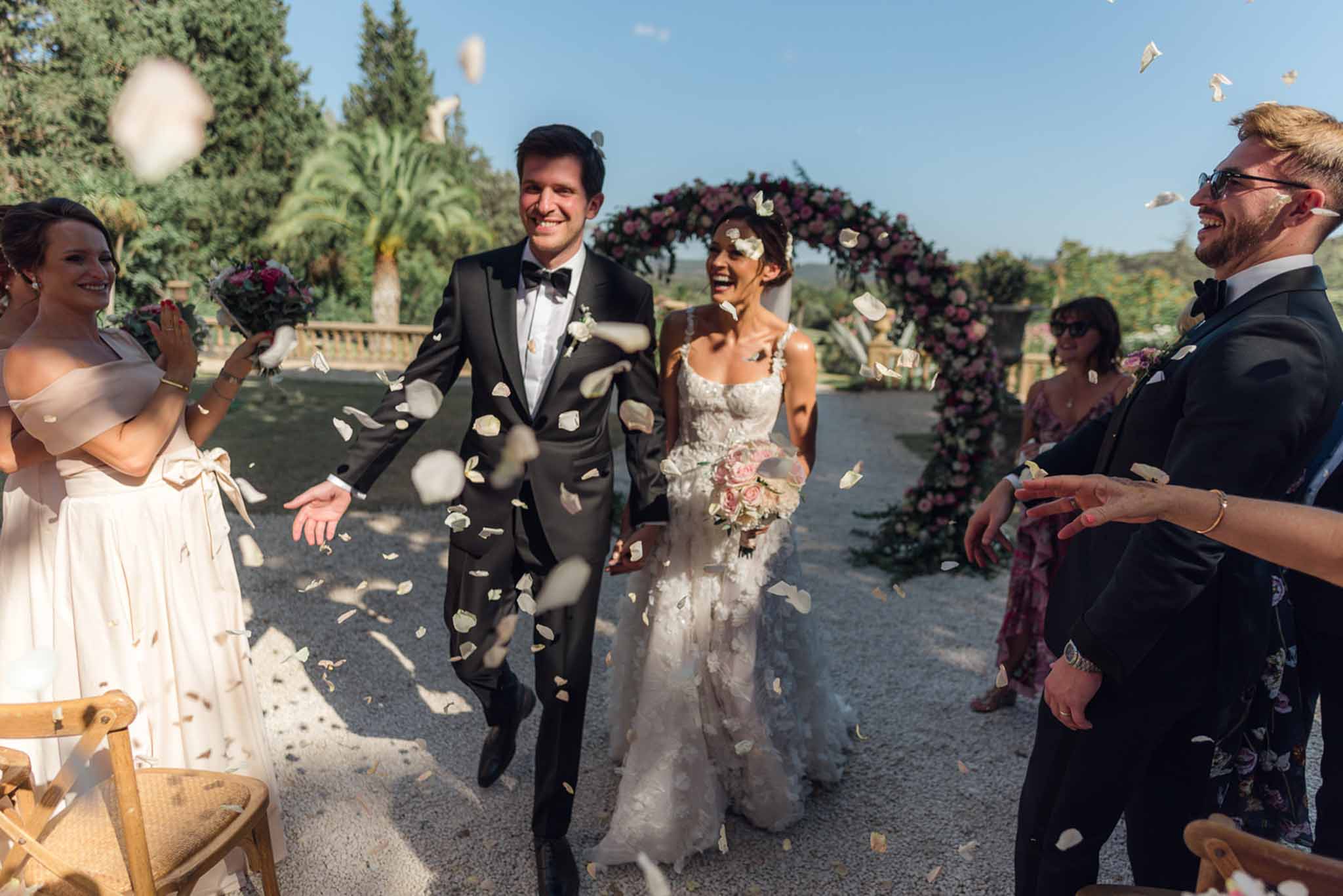Couple walks down aisle in petal toss with burgundy and blush circular rose arch behind on gravel terrace