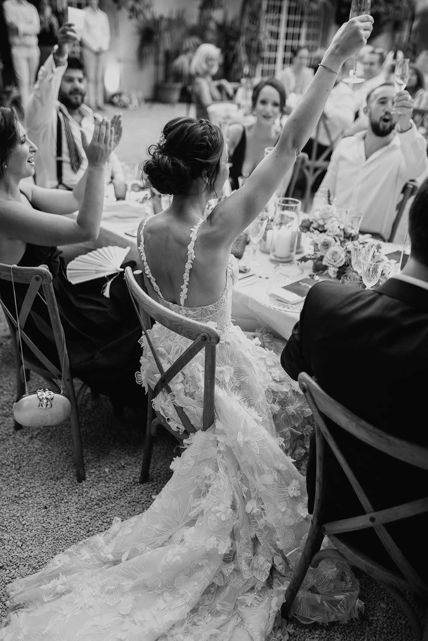 Bride from behind raising champagne glass as guests toast with raised glasses and hand fans in B&W
