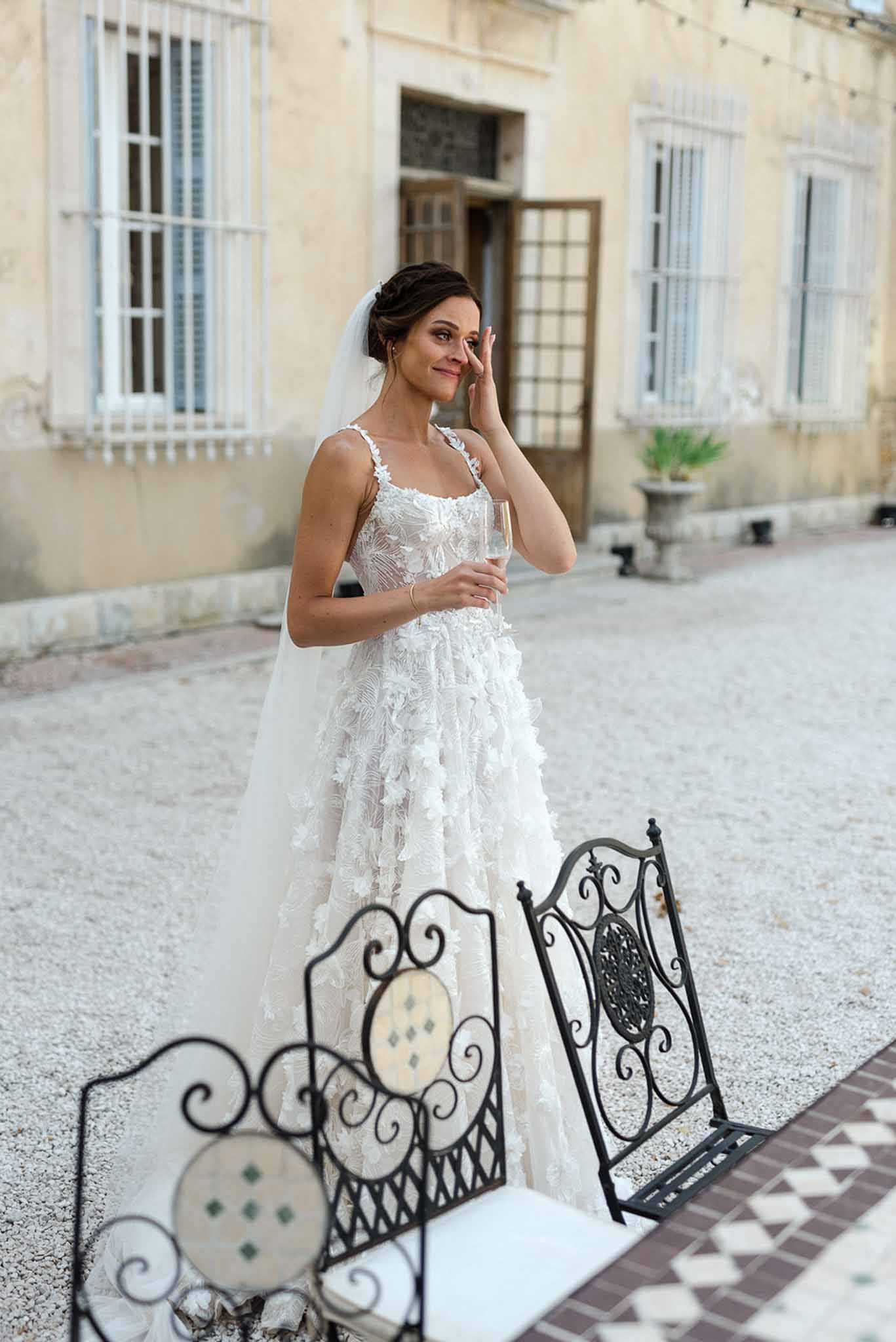 Bride in floral applique gown wiping tear while holding champagne at chateau courtyard reception
