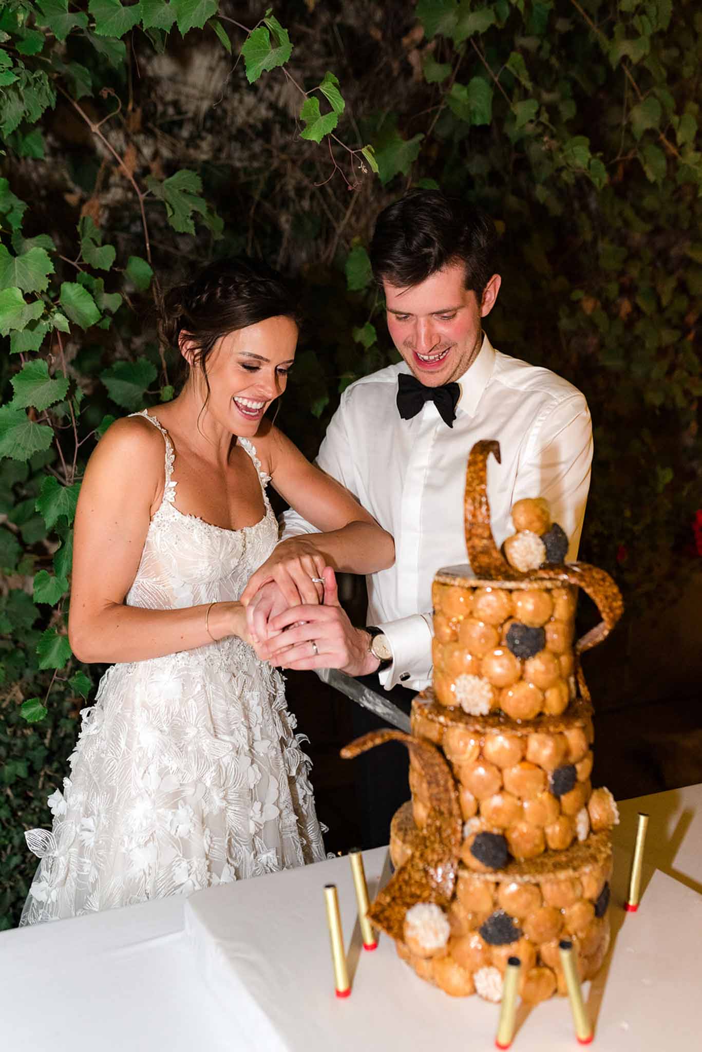 Couple laughing while cutting croquembouche tower with caramel spun sugar and gold candles at base