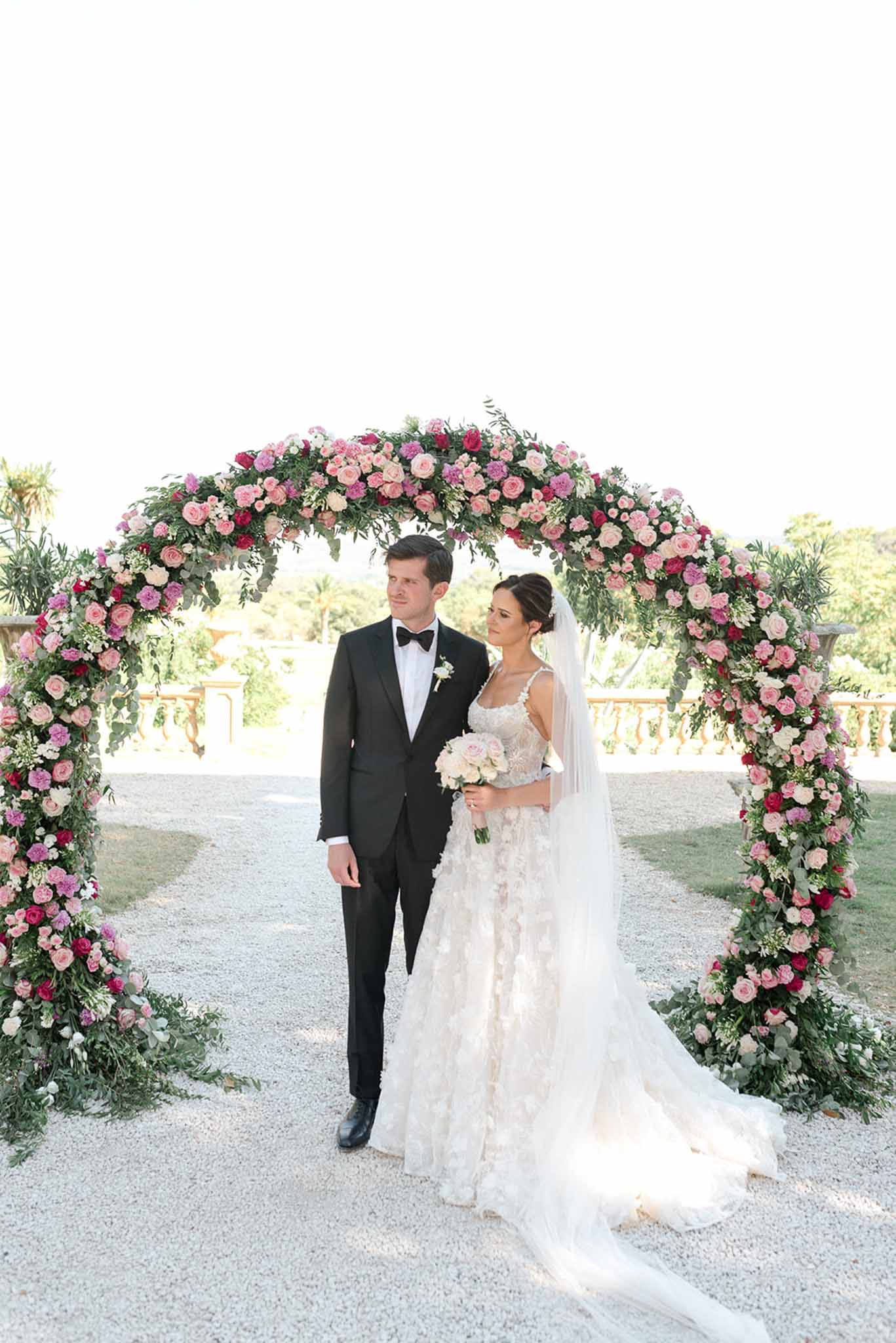 Couple posing beneath circular arch of pink and magenta roses with eucalyptus at chateau terrace