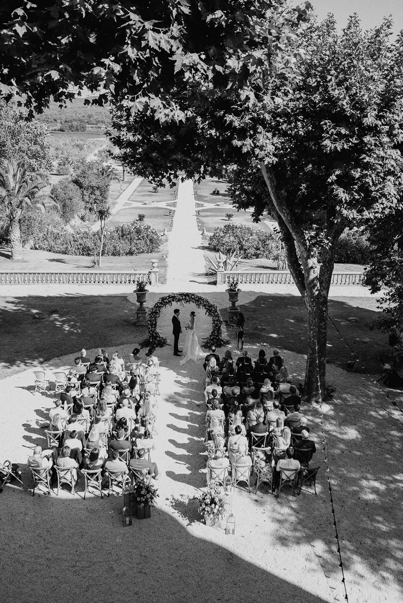 Black-and-white aerial view of outdoor chateau ceremony under circular floral arch with 80-100 seated guests in formal garden