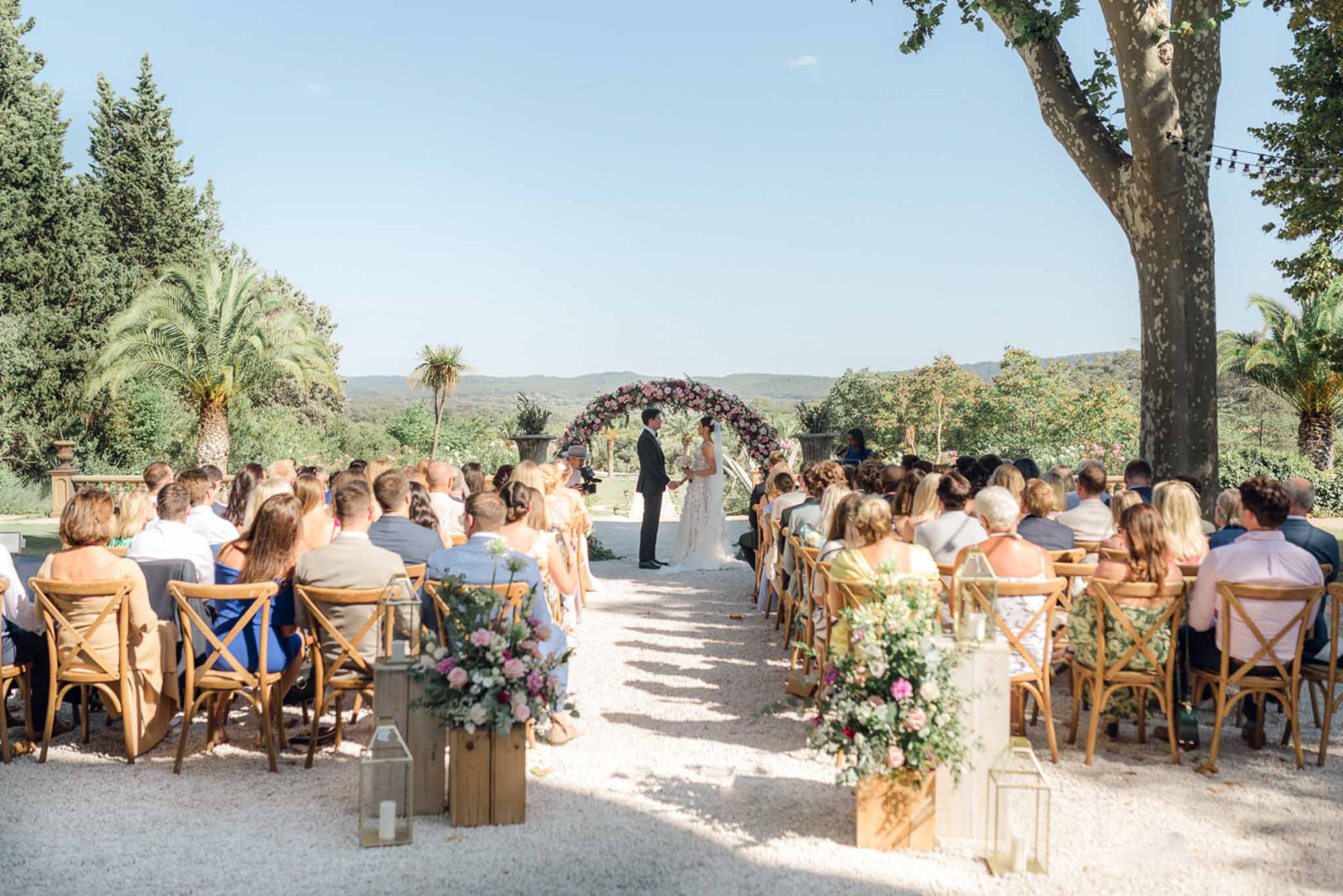 Circular blush and dusty rose floral arch with eighty guests seated on cross-back chairs overlooking rolling hills