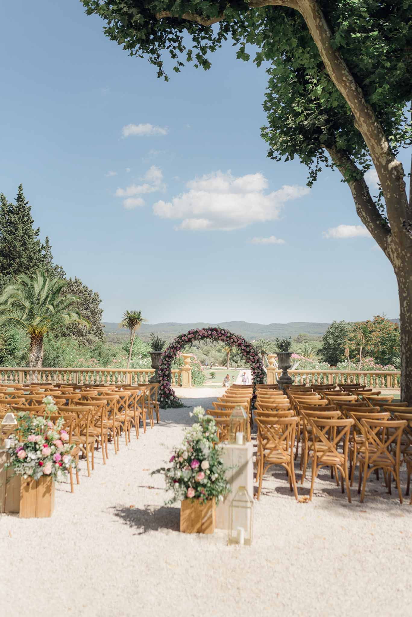 Ceremony setup with circular pink and mauve floral hoop arch, cross-back chairs, and white glass lanterns