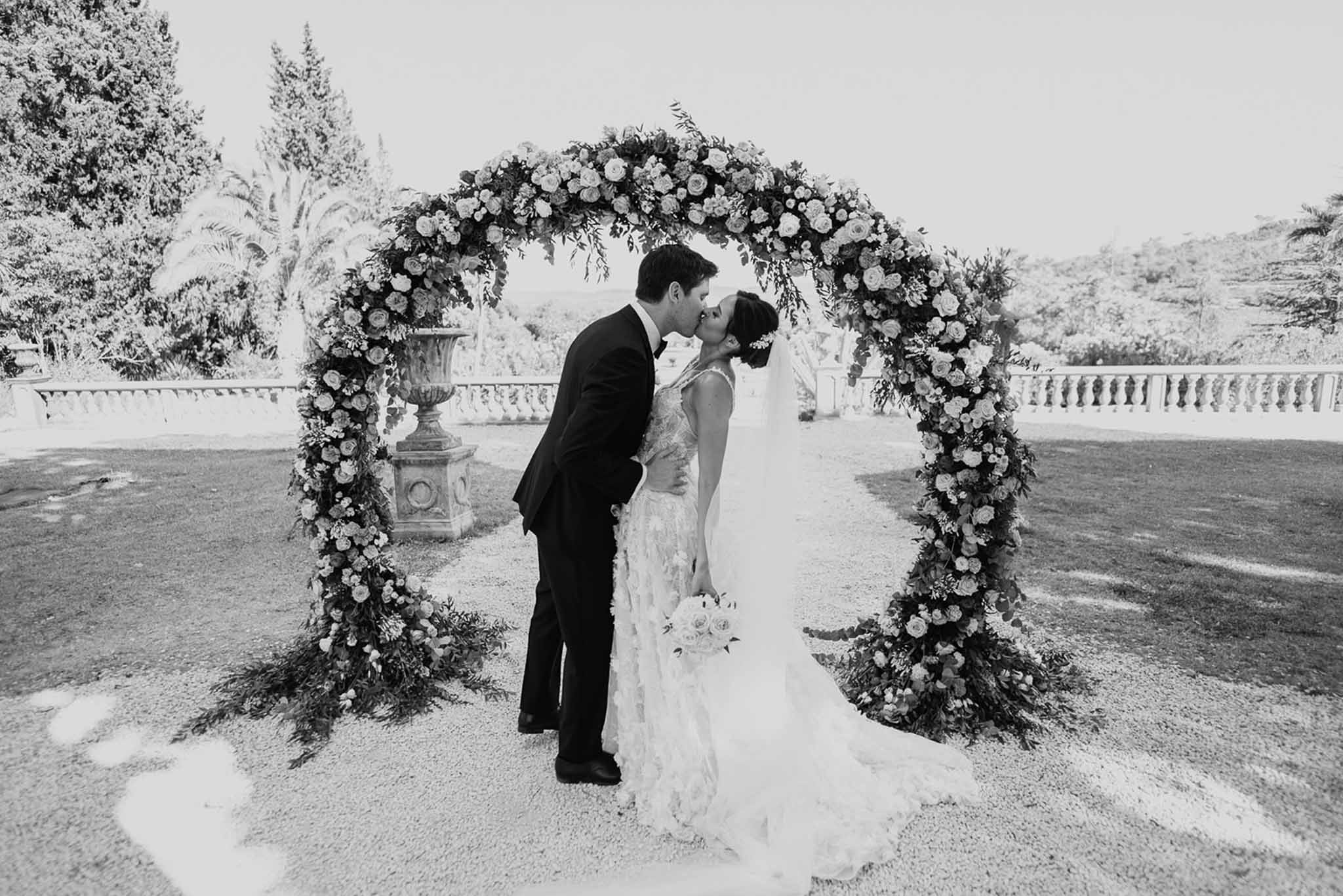 Black and white photo of bride and groom kissing under circular floral arch on chateau terrace