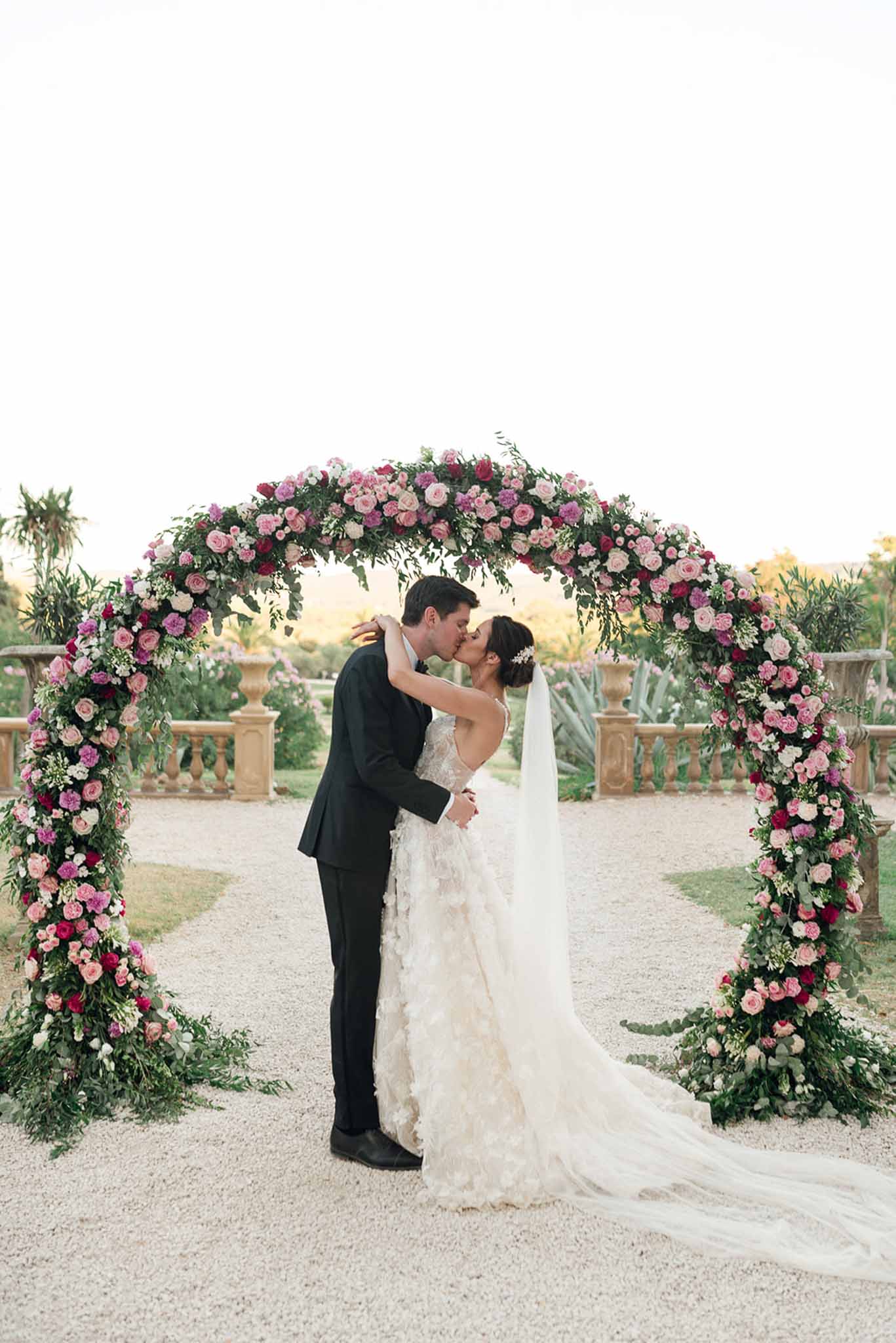 Couple kissing beneath circular arch of blush, magenta, and ivory roses in formal chateau garden