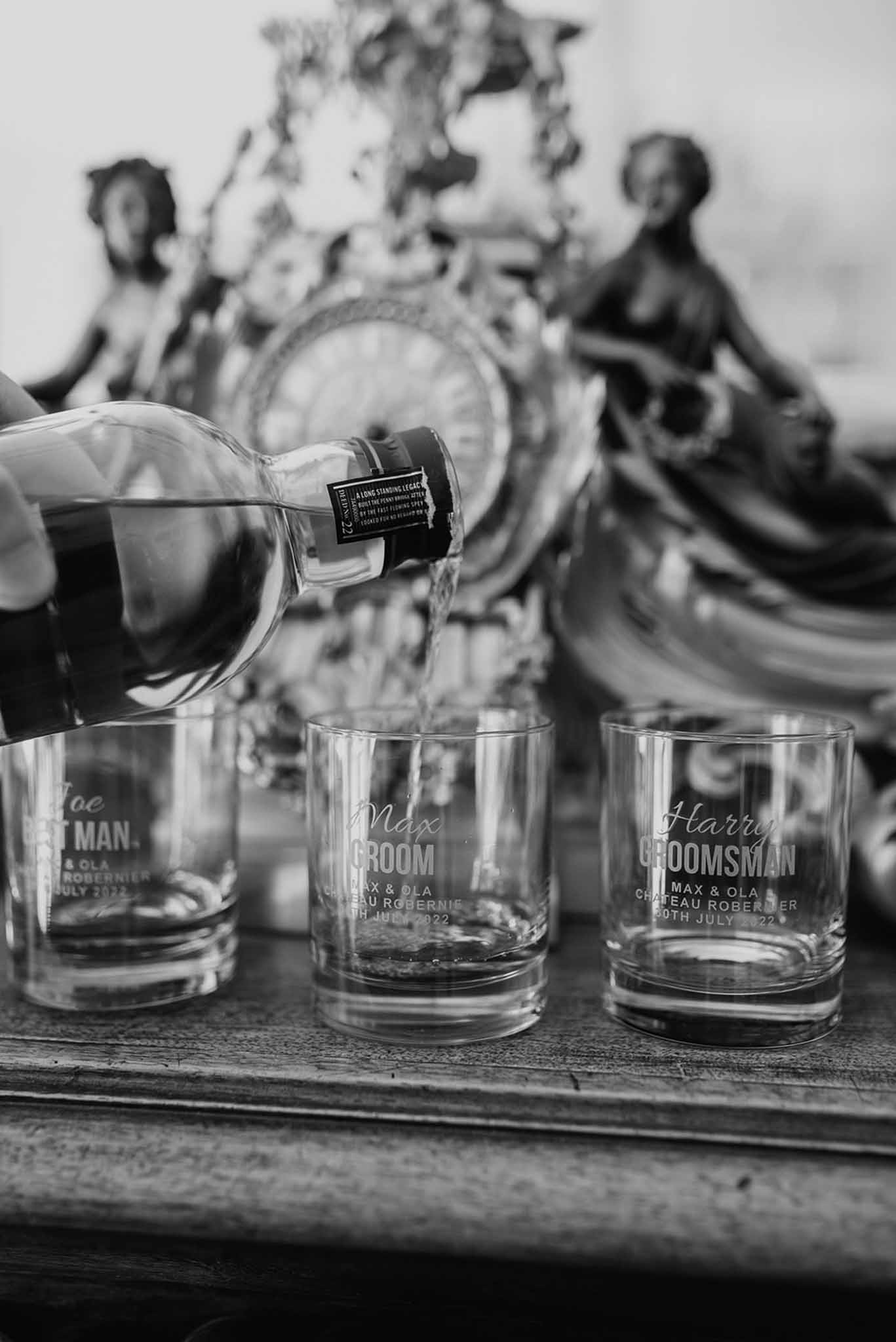 Black-and-white close-up of engraved whisky tumblers for groom and groomsmen with spirit being poured