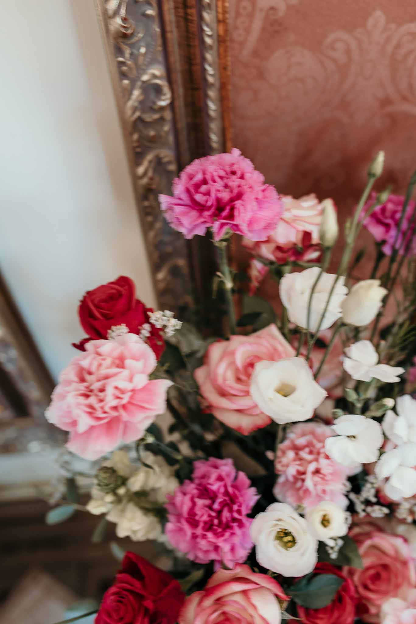 Pink carnations, blush roses, and red roses arranged against gilded mirror and damask wallpaper