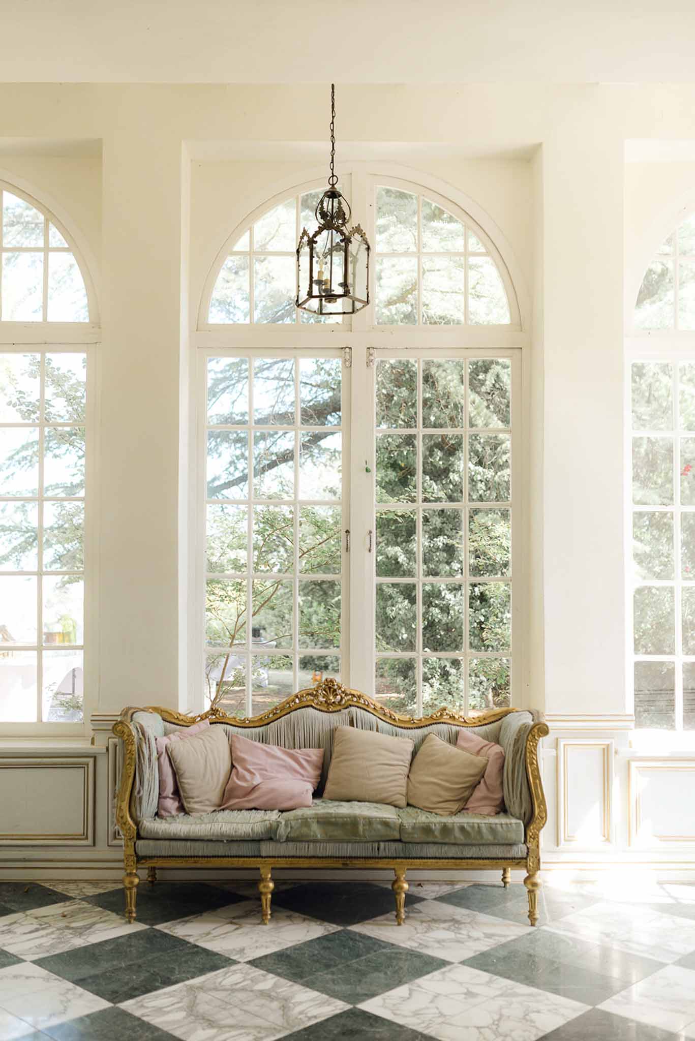 Chateau salon with gilded sage green sofa, black and white marble floor, and arched French doors
