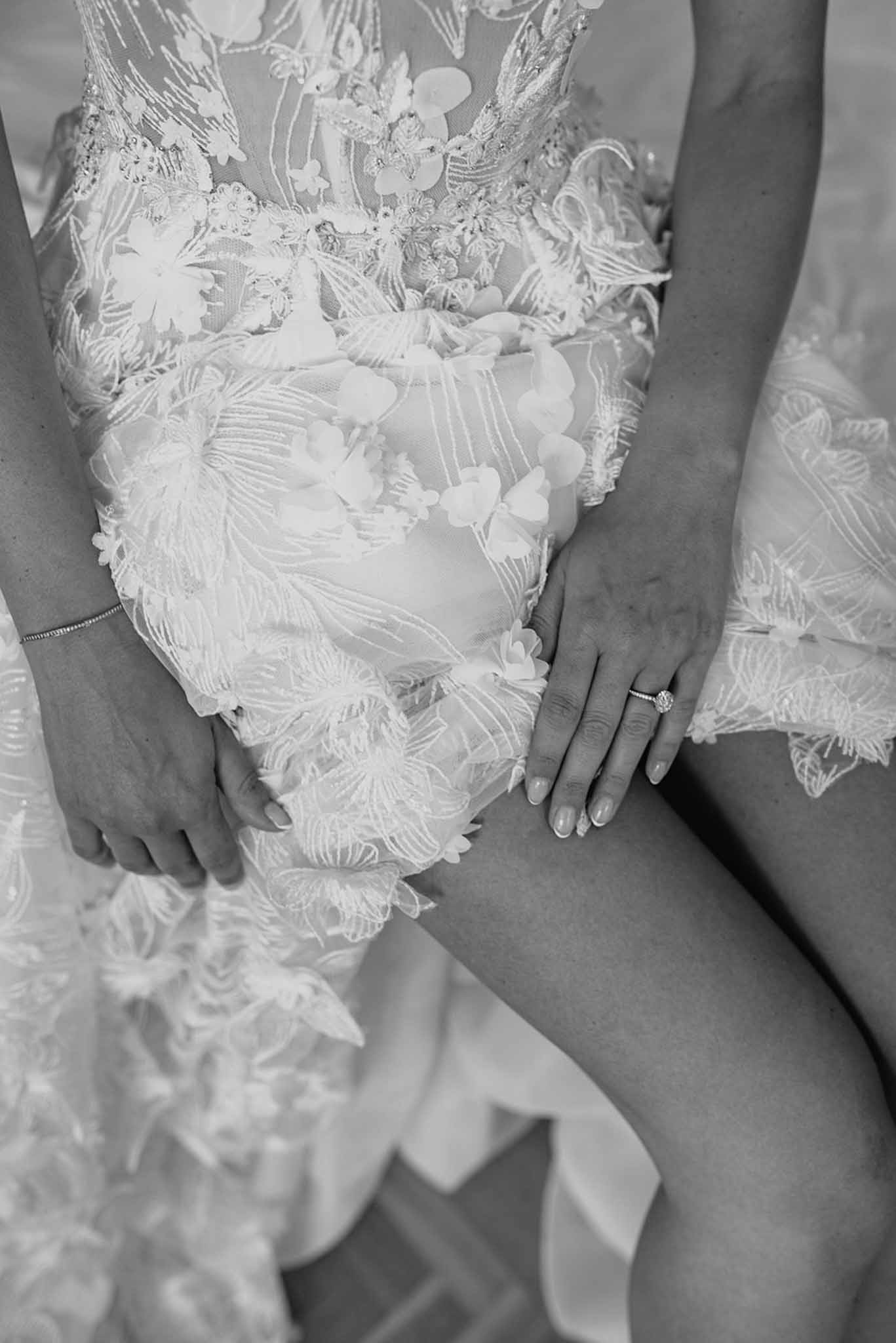 Black and white close-up of bride's hands with solitaire diamond ring on embellished 3D floral lace gown