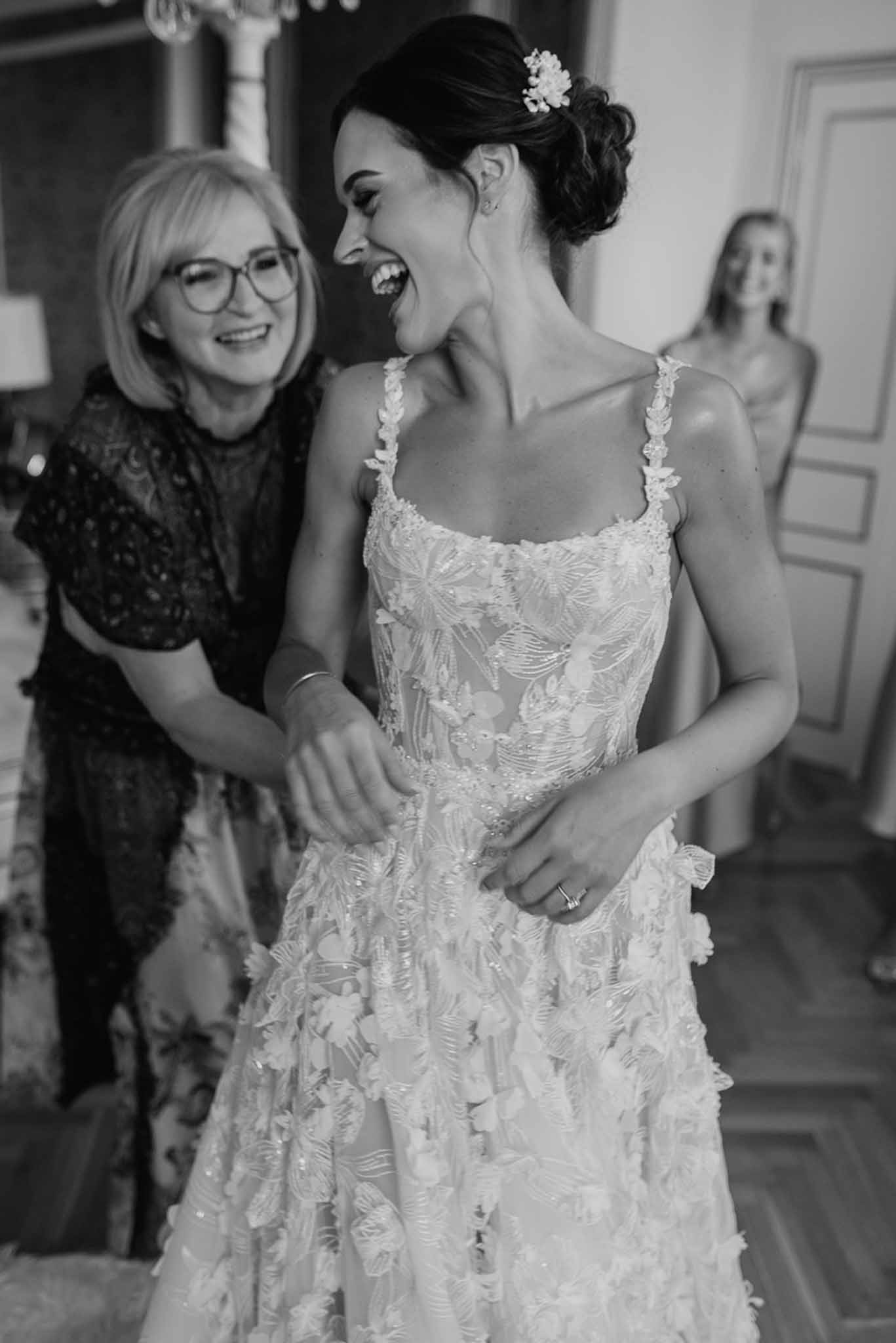 Black-and-white candid of bride laughing as older woman fastens her floral applique gown in a chateau room