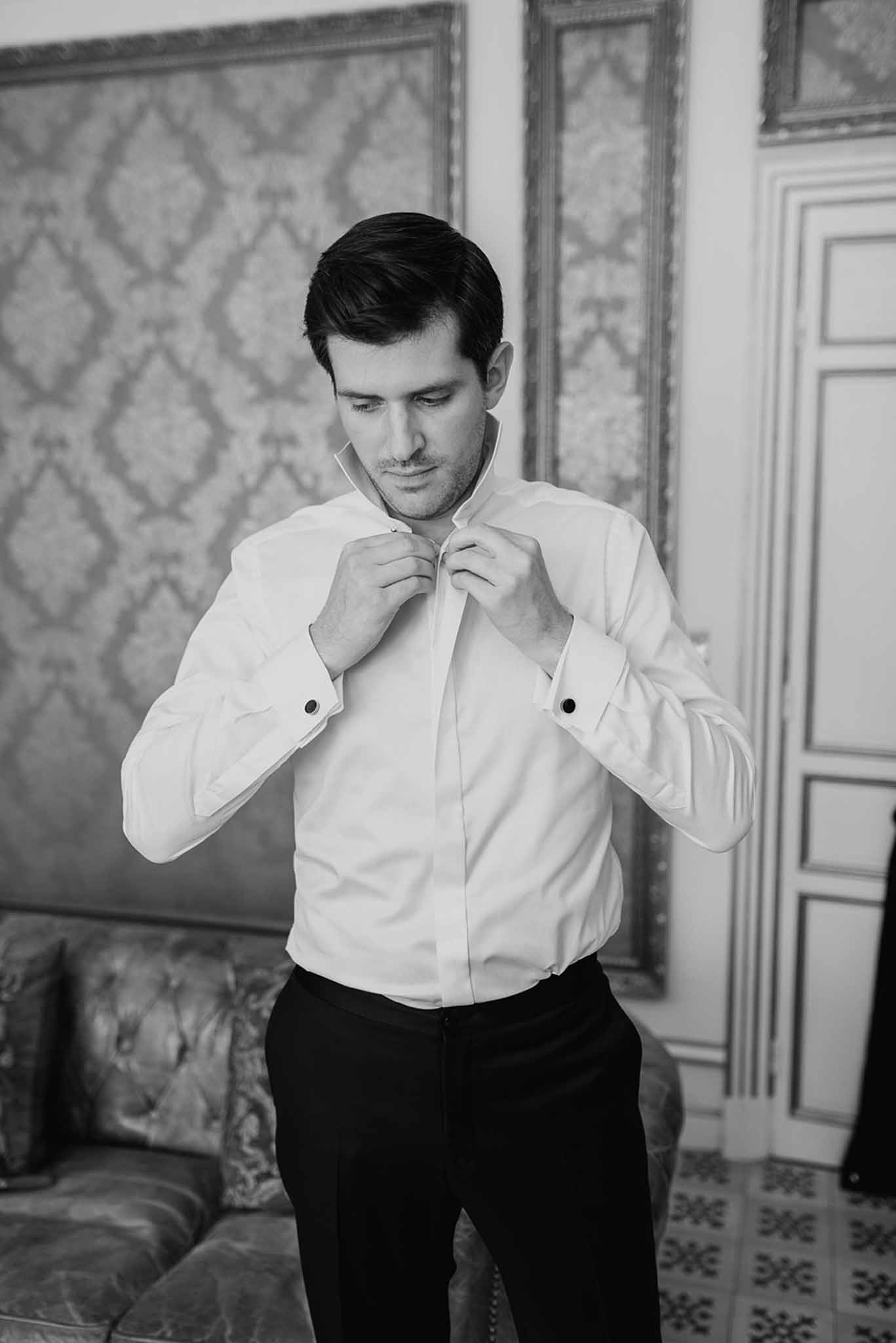 Black and white groom buttoning white shirt in ornate chateau interior with tufted sofa and moulded door frame