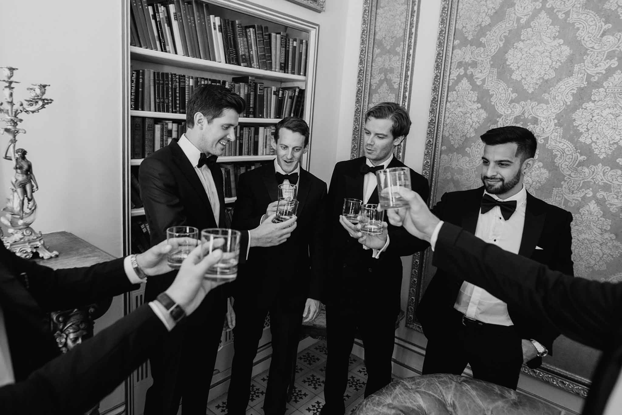 Groomsmen in black tuxedos sharing a whisky toast in a chateau room with damask wallpaper and bookshelf