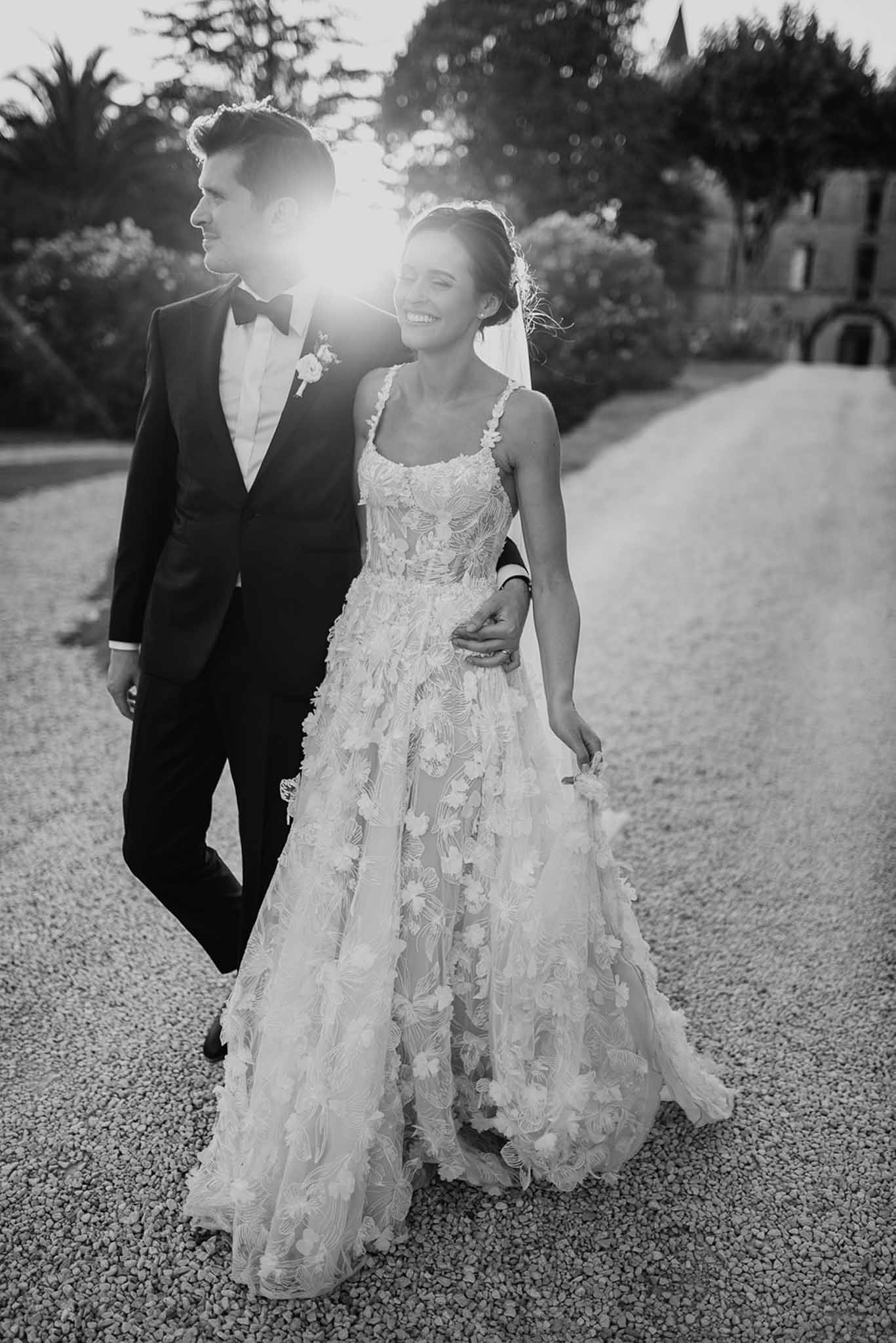 Black-and-white portrait of laughing bride and groom walking arm in arm on chateau driveway with sun flare