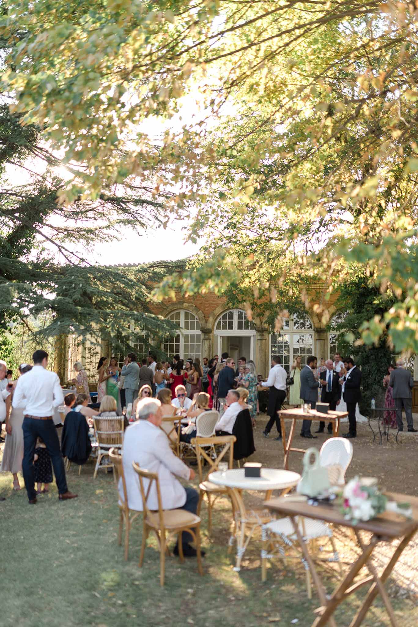 Fifty guests mingle at cross-back chairs and bistro tables under trees at red brick chateau terrace