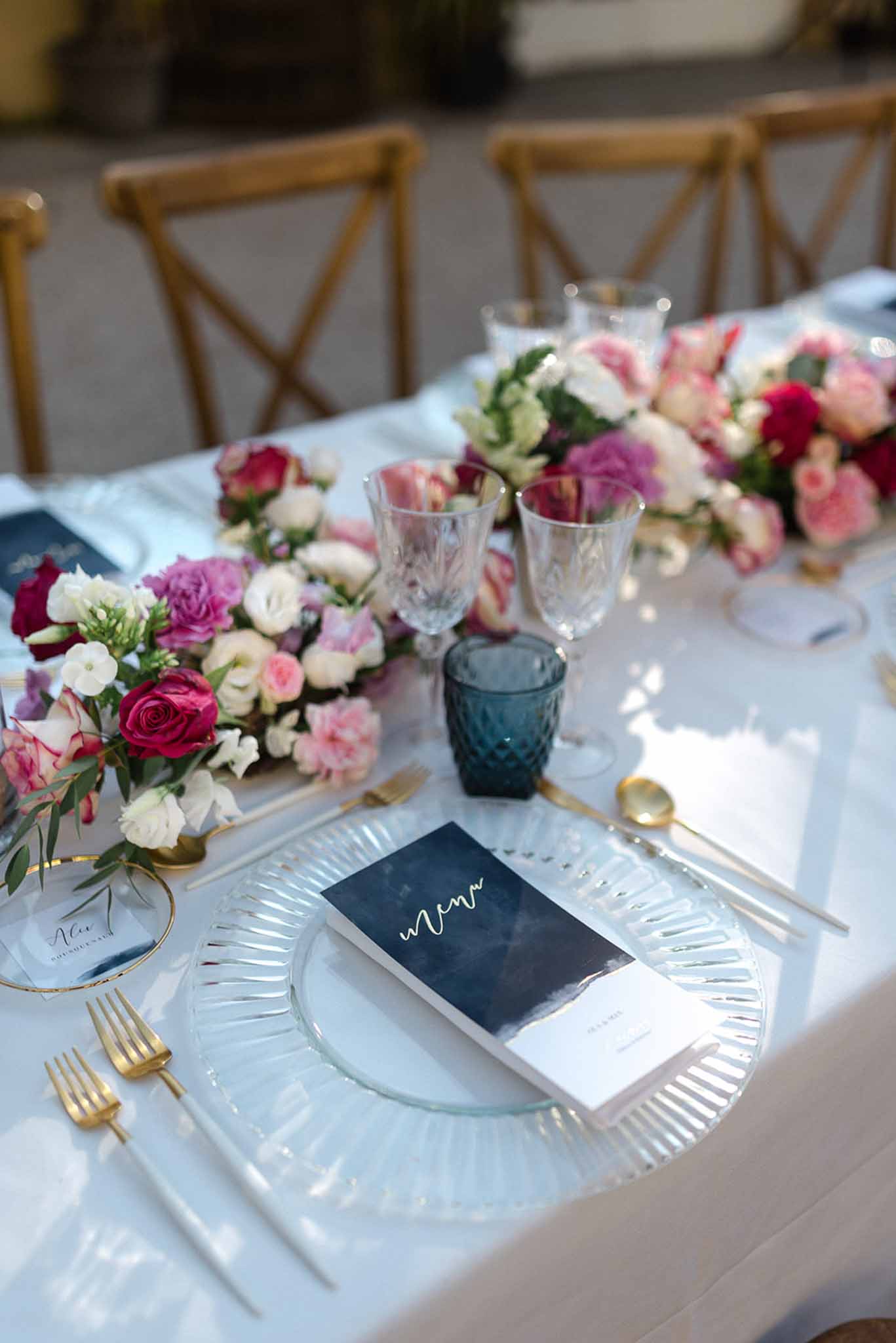 Reception place setting with ribbed glass charger, gold flatware, navy menu, and crimson rose runner