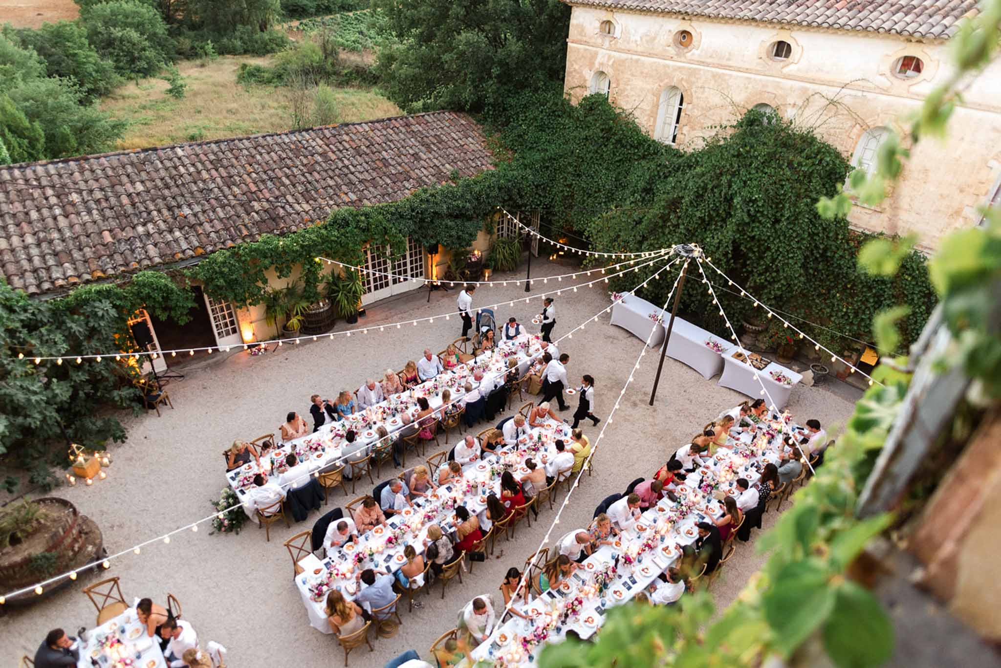 Aerial view of outdoor reception dinner with 80-100 guests at long banquet tables in stone courtyard