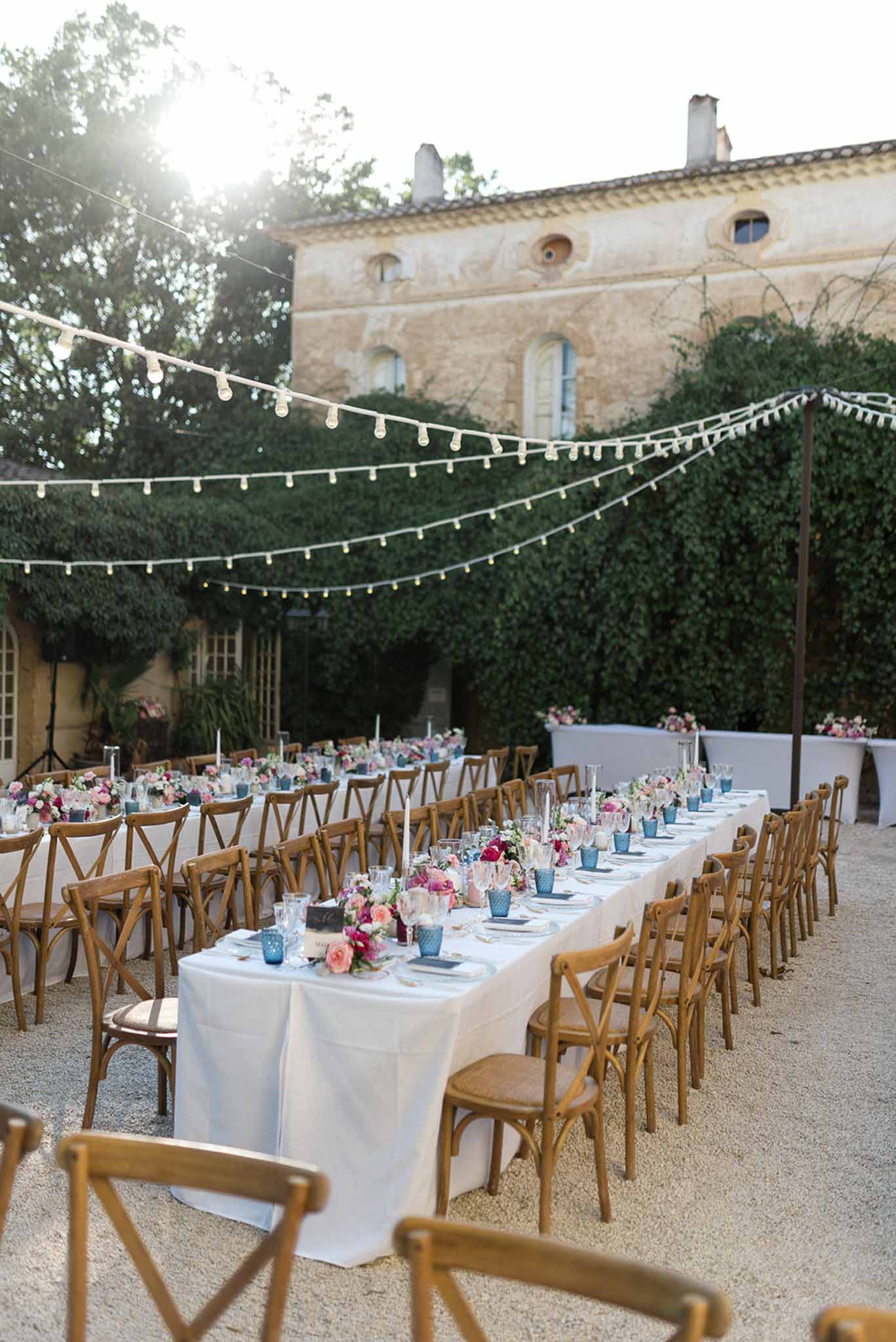 Banquet tables with pink and magenta florals, dusty blue glass, and festoon lights at ivy-walled courtyard