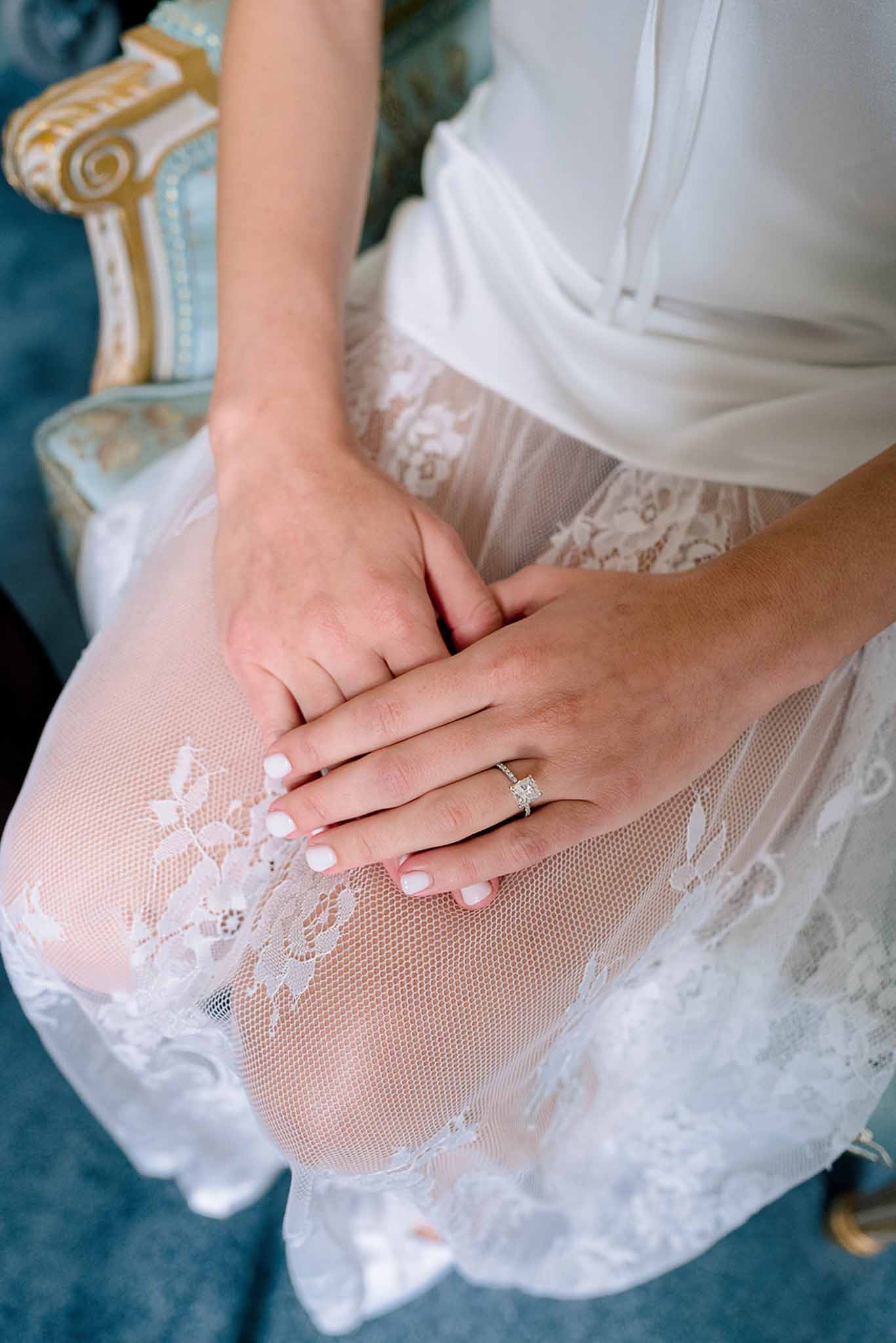 Close-up of bride's hands showing engagement ring, lace garter, and white bridal robe while seated