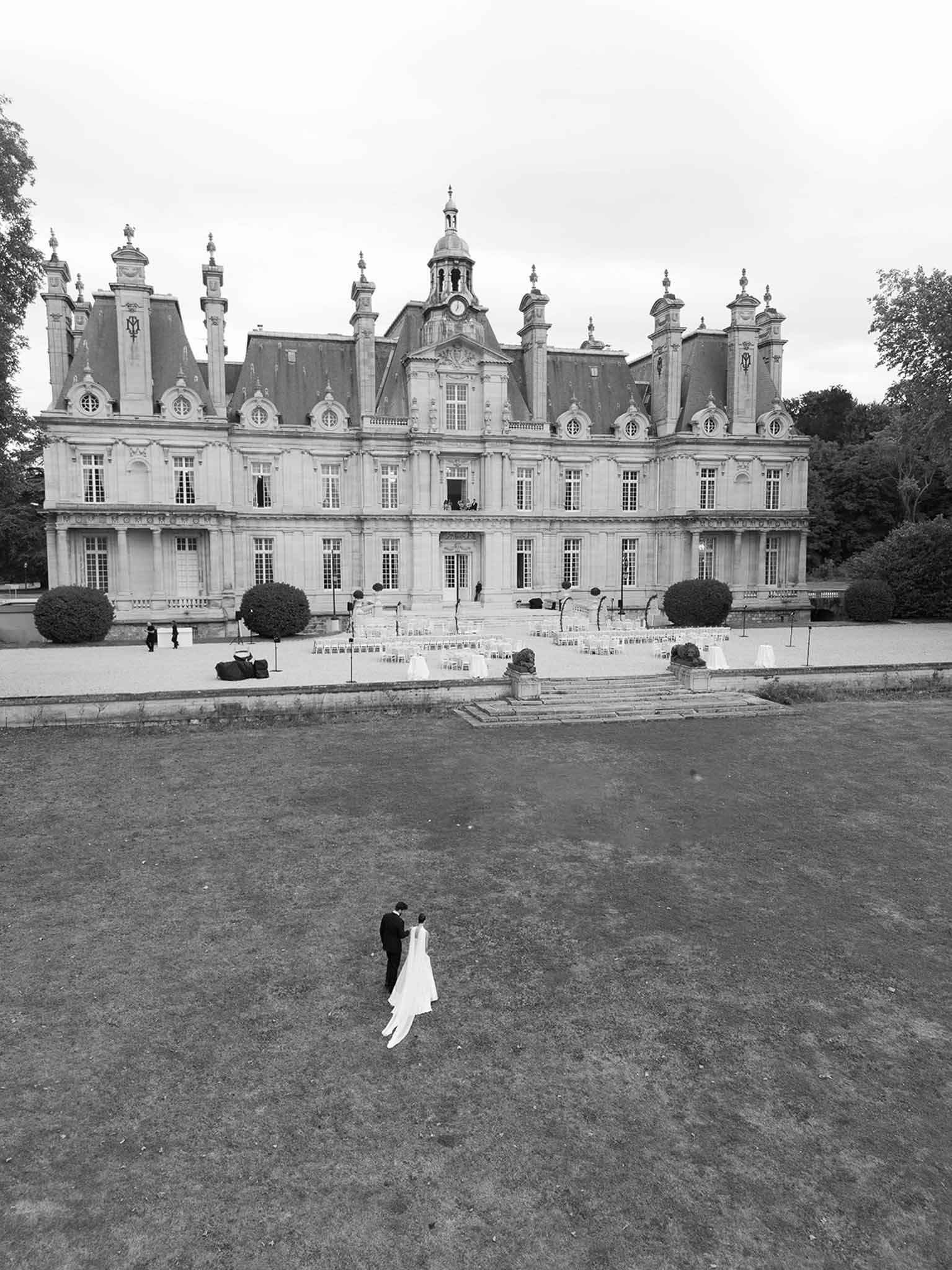 Black and white aerial view of couple on lawn before grand chateau with clock tower and ceremony chairs