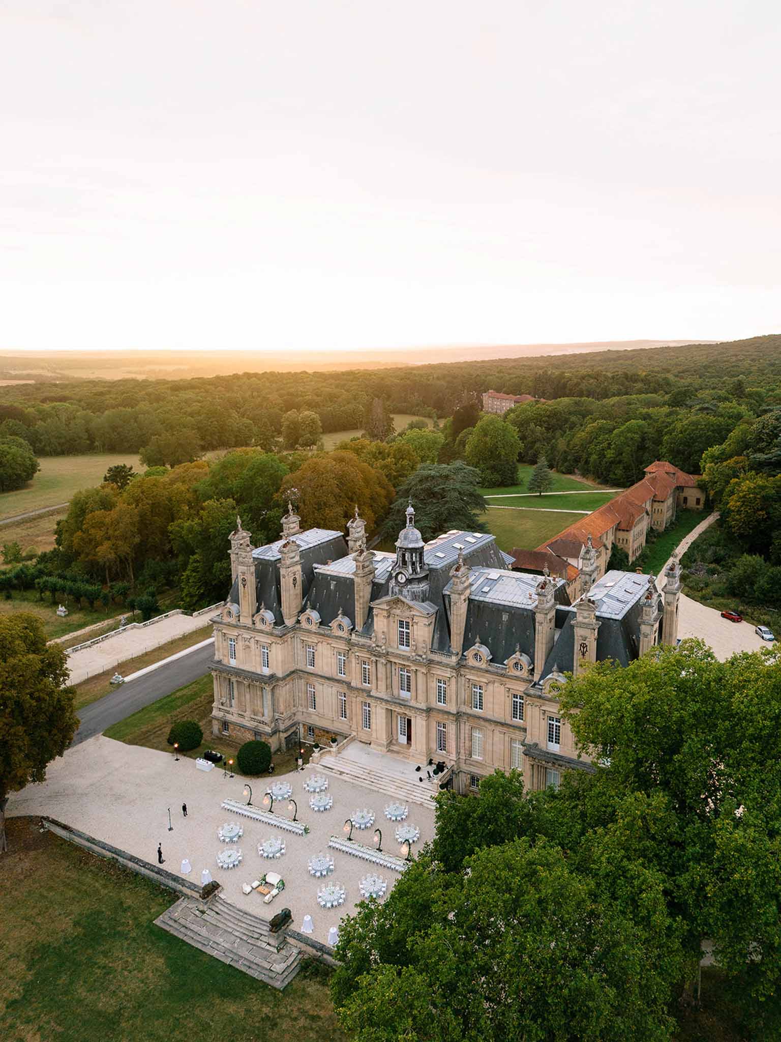 Aerial dusk view of classical chateau with mansard roof and reception tables in gravel courtyard