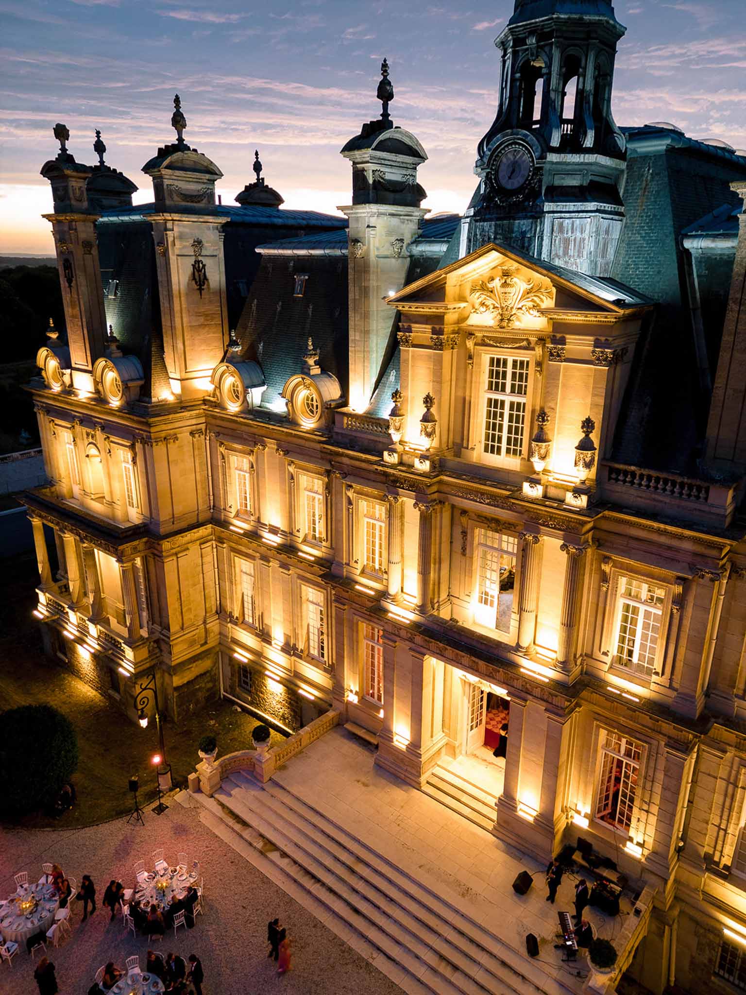 Aerial dusk view of illuminated French chateau with outdoor reception tables and guests on terrace below