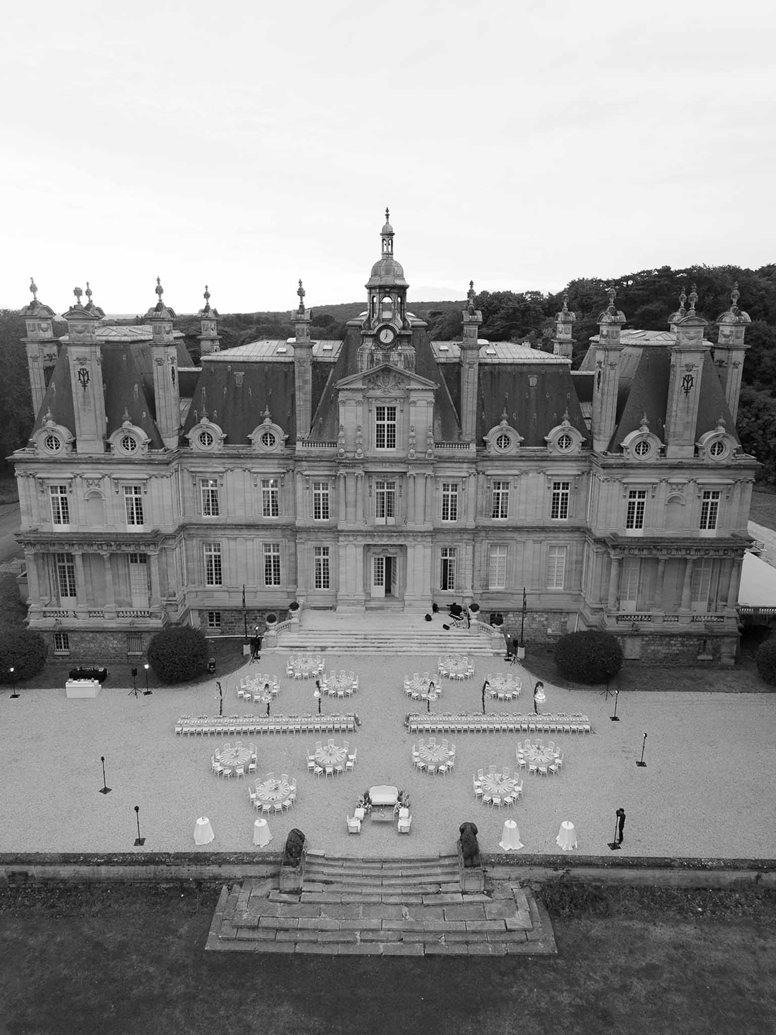 Black and white aerial of grand chateau with clock tower and reception tables on gravel forecourt