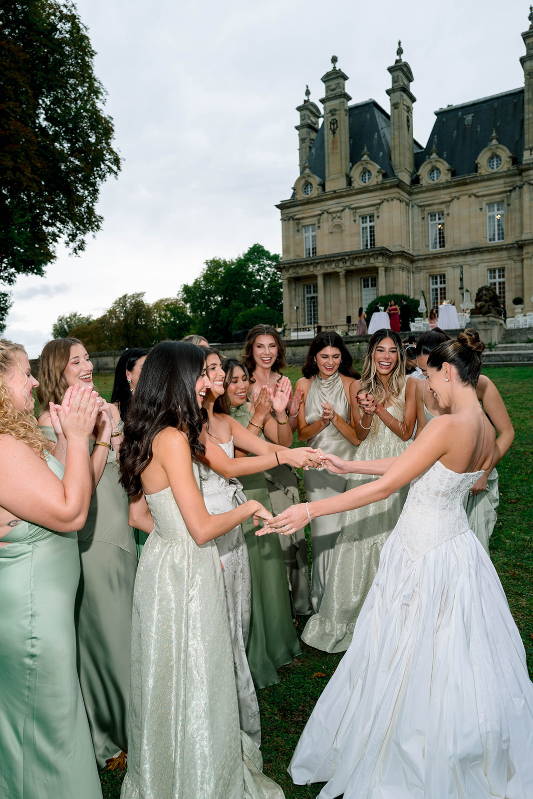 Bride surrounded by ten bridesmaids in sage and champagne dresses admiring ring on chateau lawn