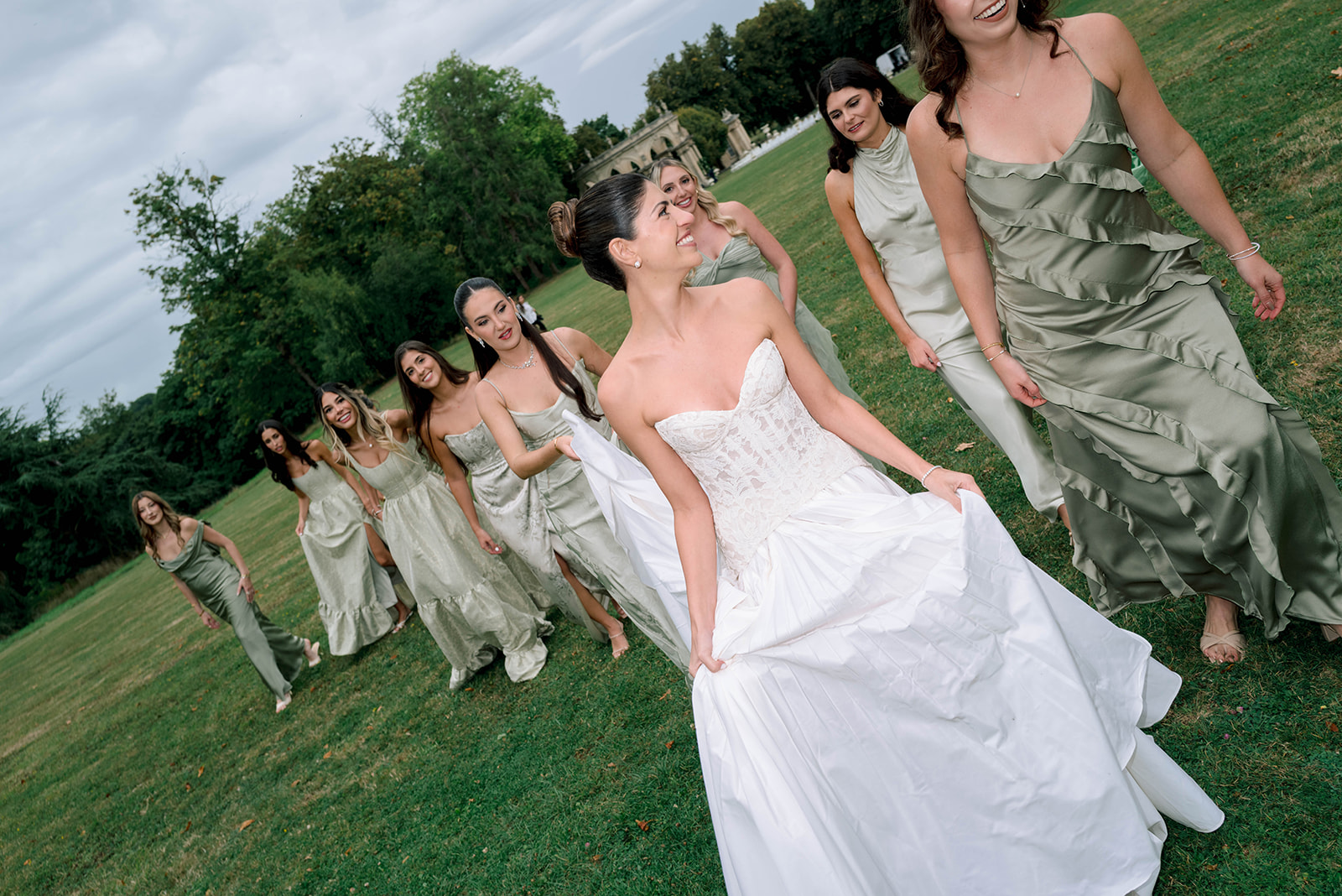 Bride in strapless lace ballgown walking on a lawn with seven bridesmaids in mismatched sage green dresses