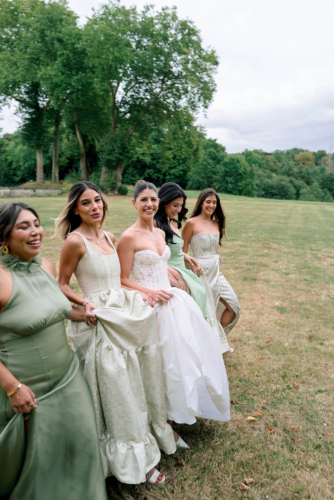 Bride with five bridesmaids in sage green and silver-grey gowns walking across lawn