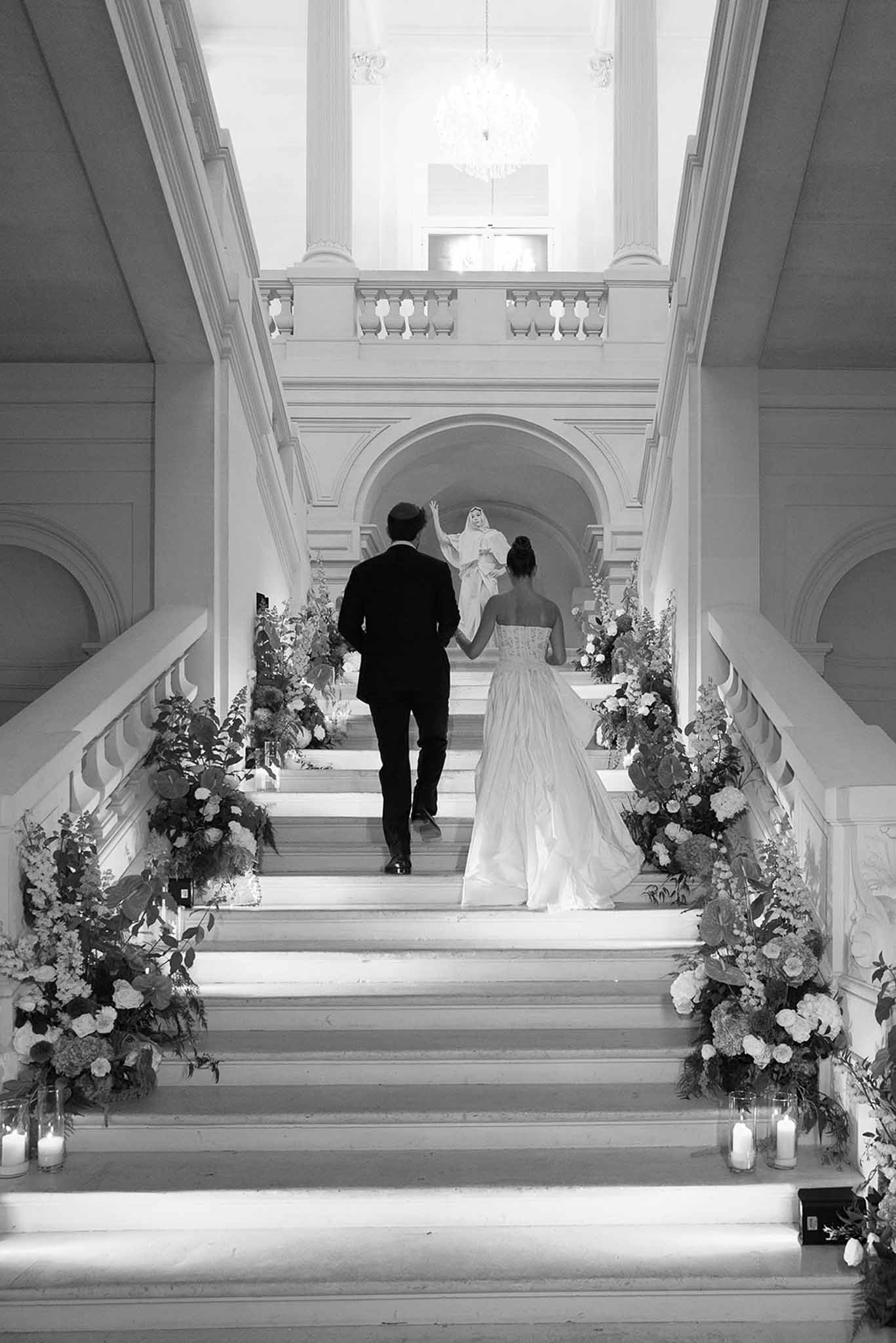 Black and white of couple ascending grand staircase lined with floral arrangements and hurricane candles
