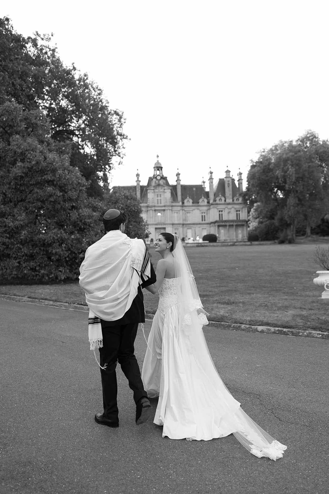 Black and white photo of bride and groom walking toward grand French chateau, bride looking back smiling