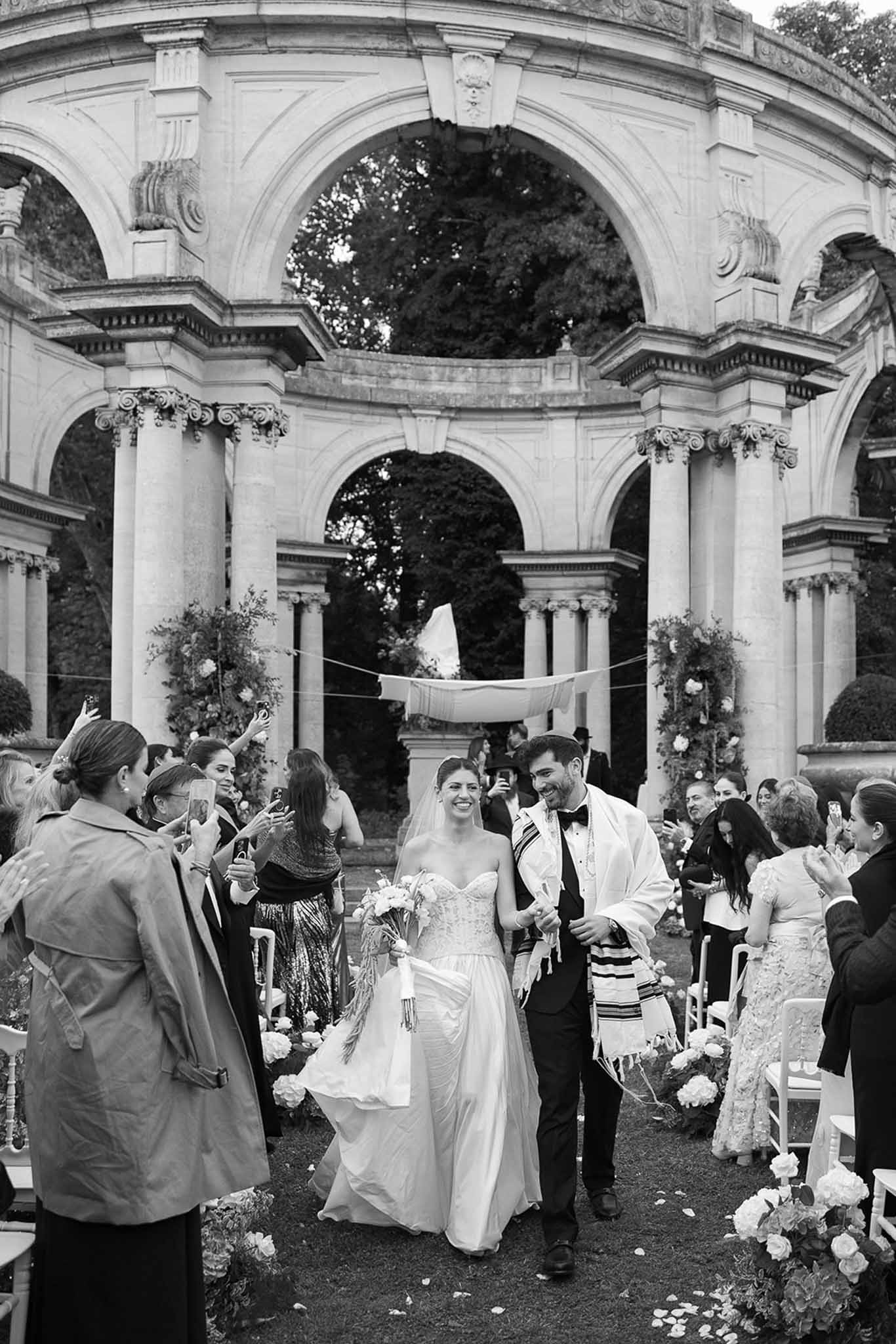 Black-and-white recessional after Jewish ceremony under neoclassical colonnade with guests and chuppah behind
