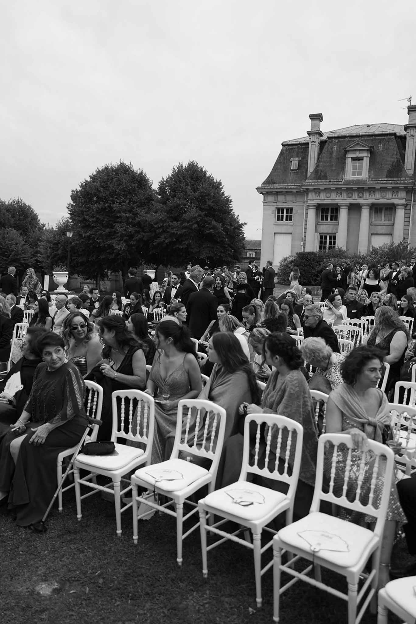 Black and white elevated shot of 80 guests in Napoleon chairs at ceremony before chateau with columns