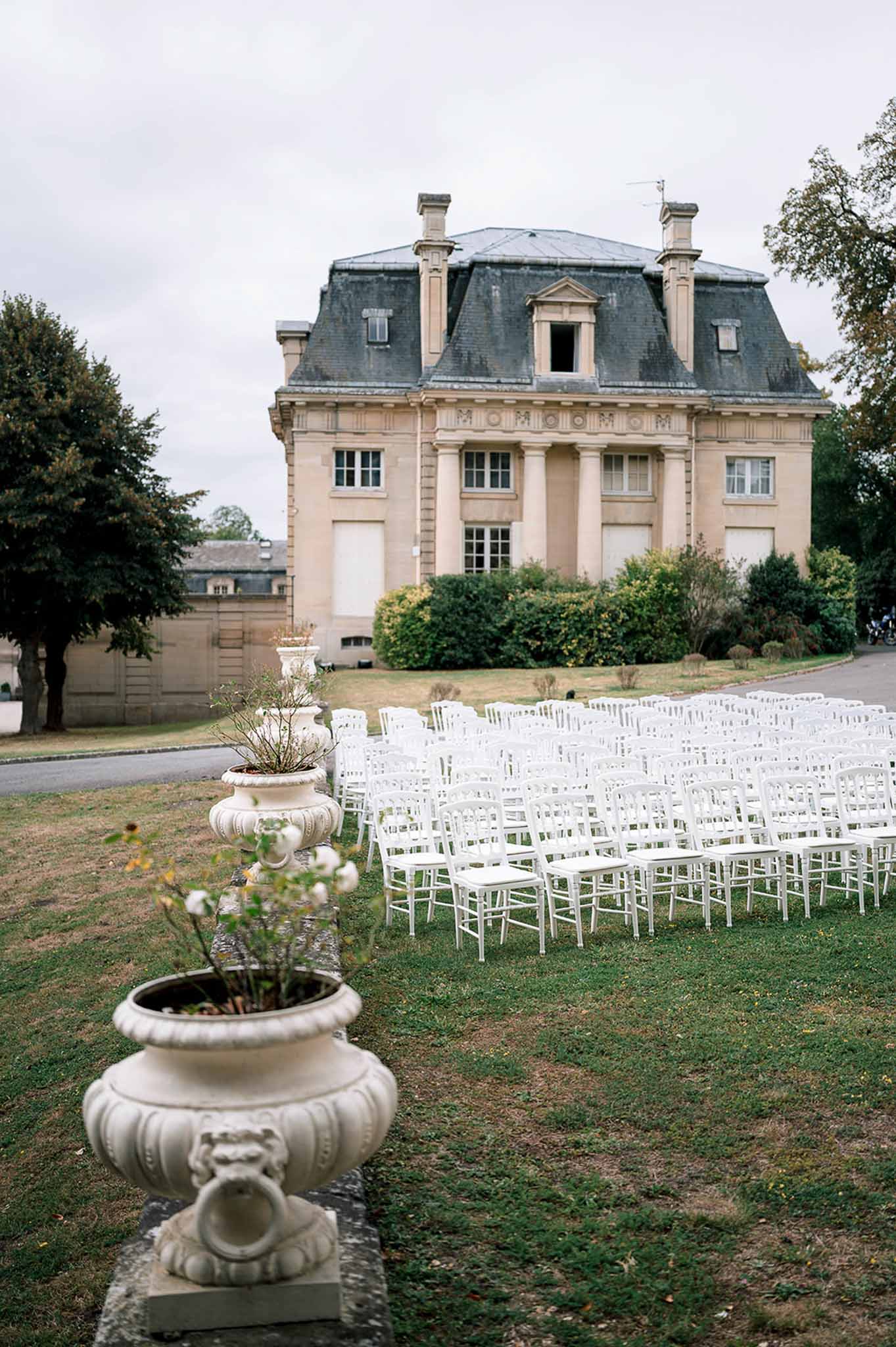 French chateau ceremony setup with white chairs on lawn, stone urns with lion-head details in foreground
