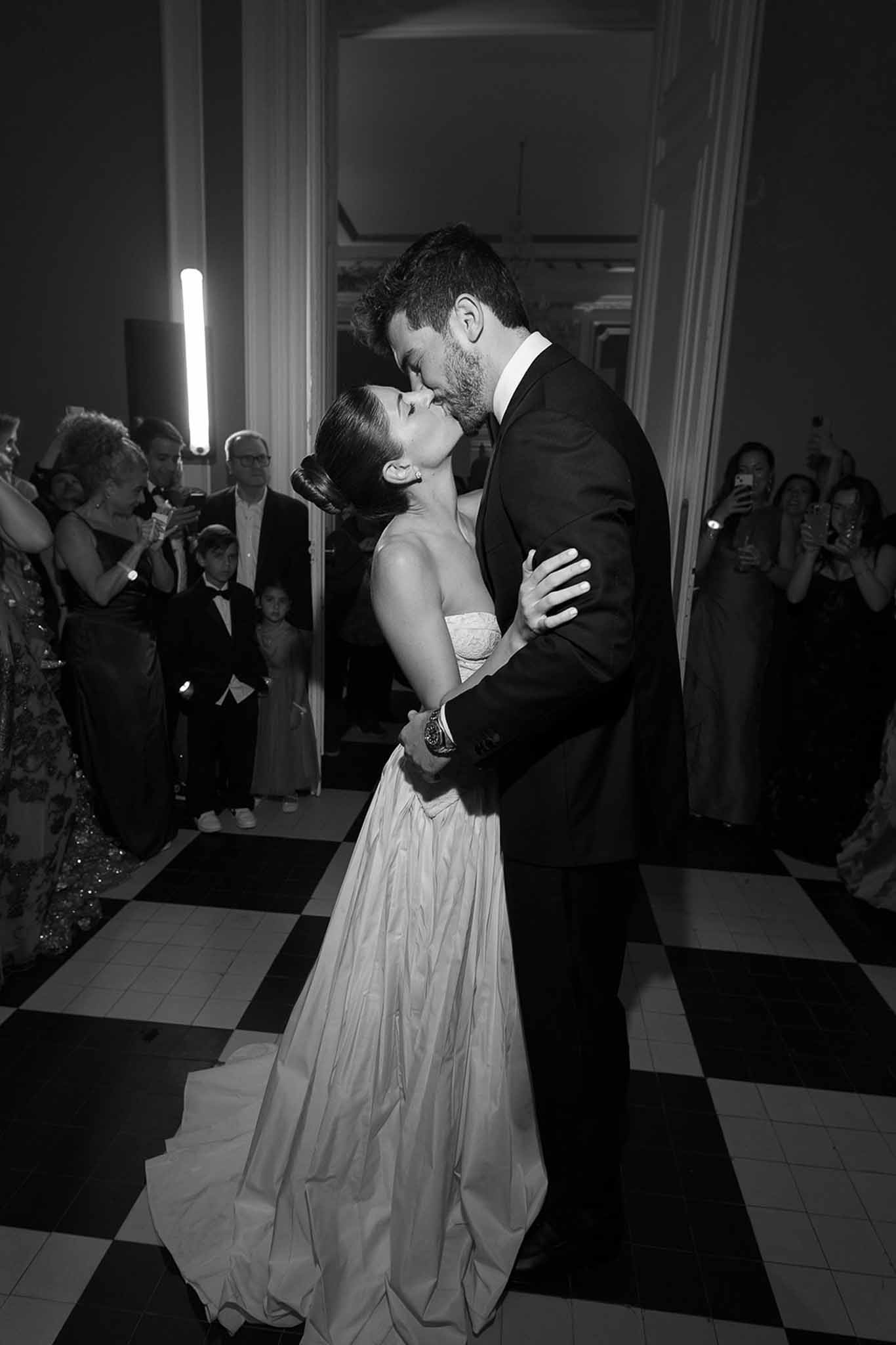 Black and white photo of bride and groom kissing during first dance on checkered floor surrounded by guests