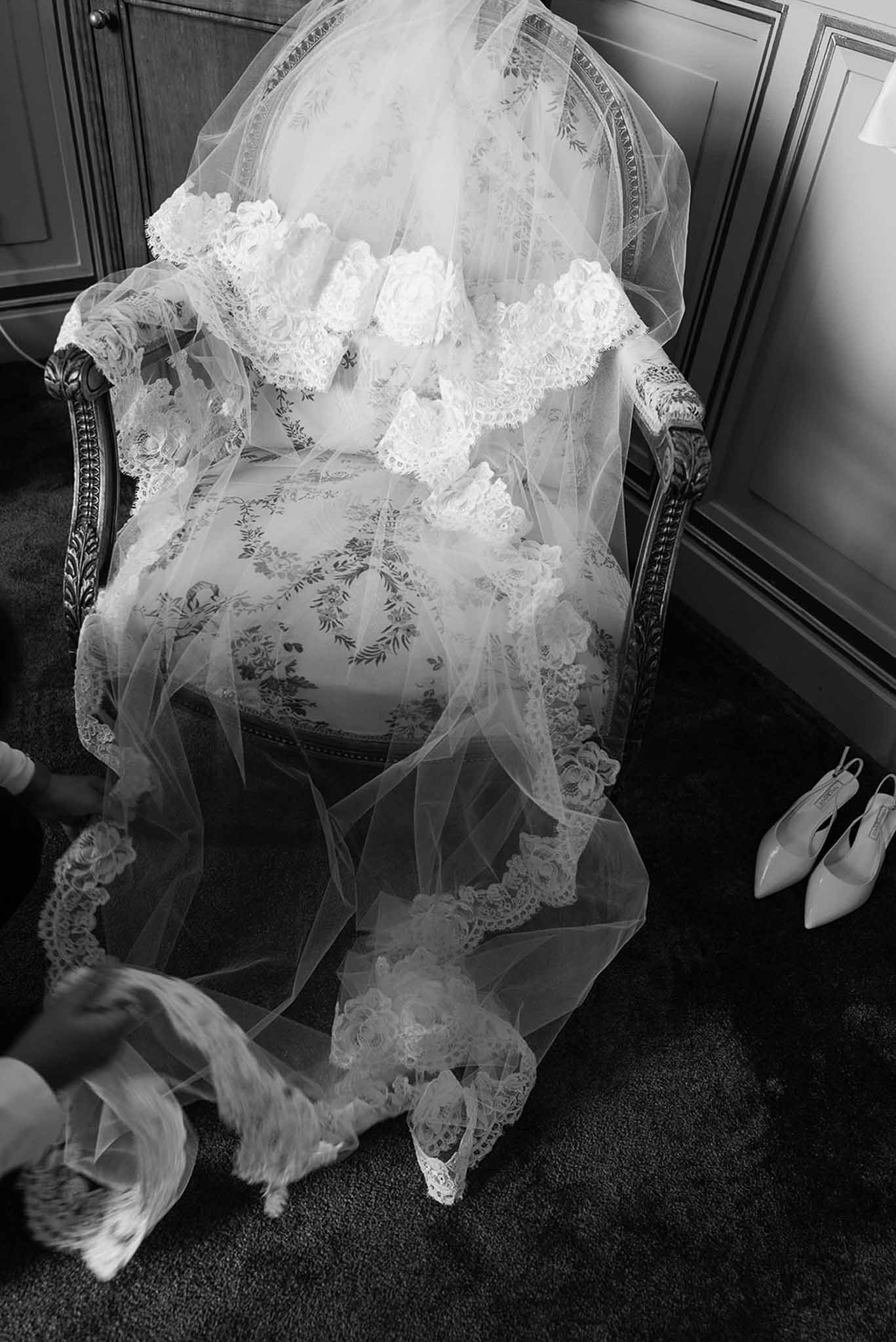 Black-and-white detail of bridal veil with lace trim draped over ornate armchair beside white satin slingback heels