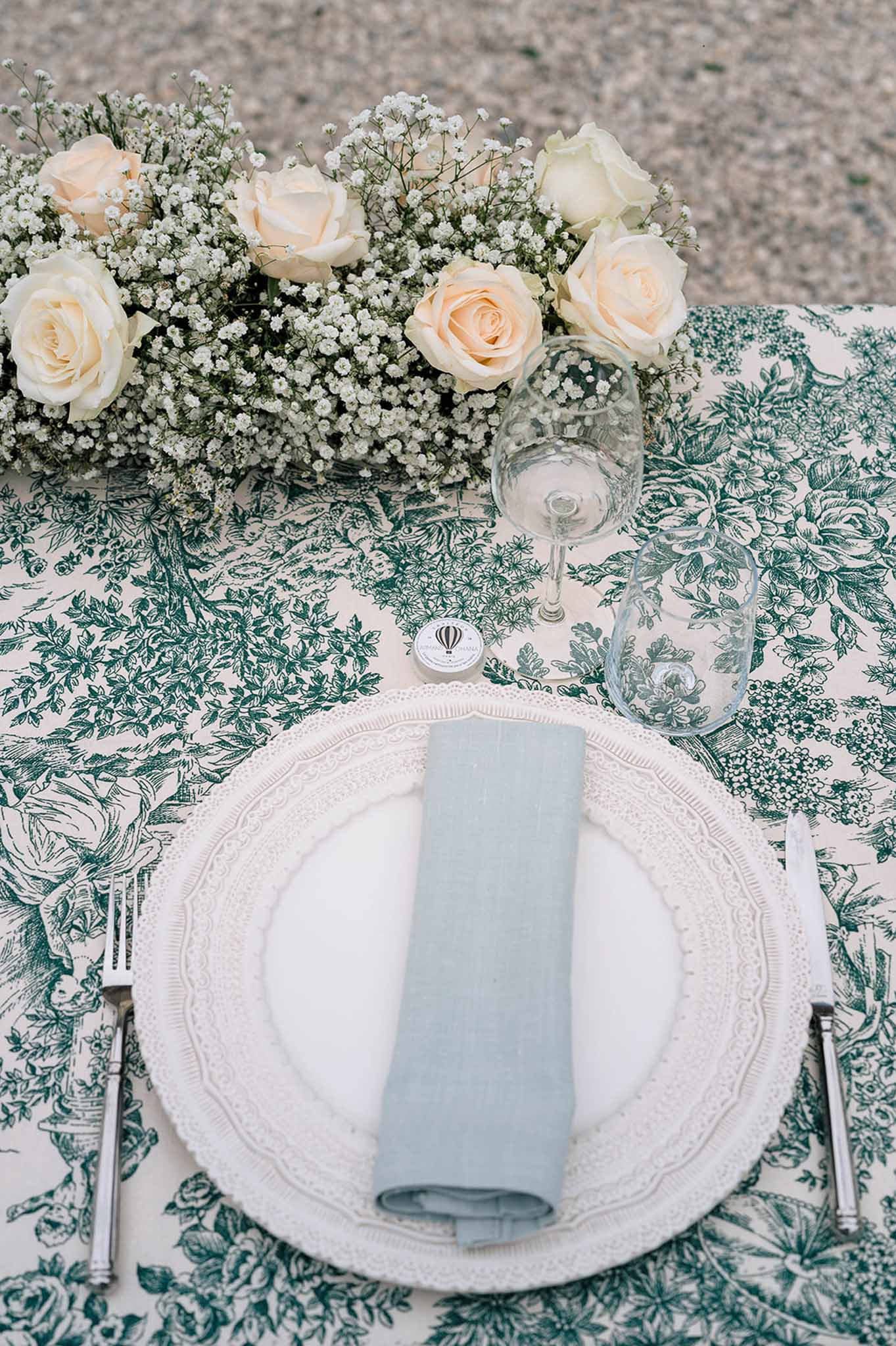 Place setting with dusty blue napkin on toile de Jouy cloth with ivory roses and baby's breath centerpiece