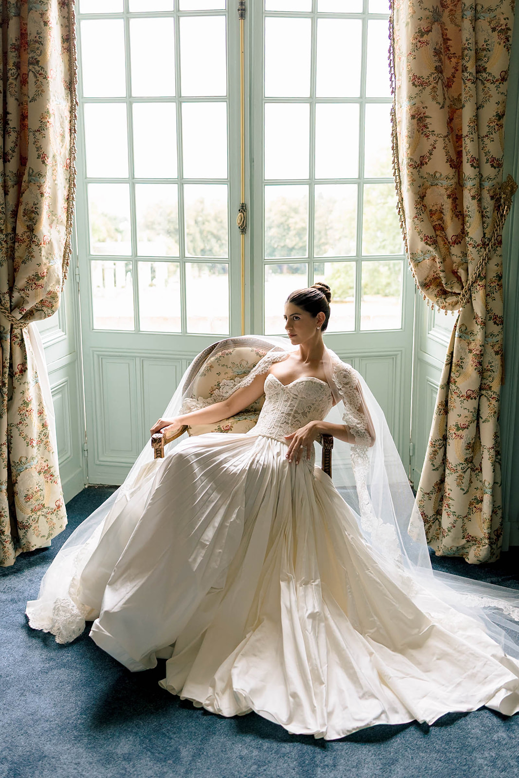 Bride seated in armchair wearing ivory lace corset ball gown with cathedral veil inside chateau room