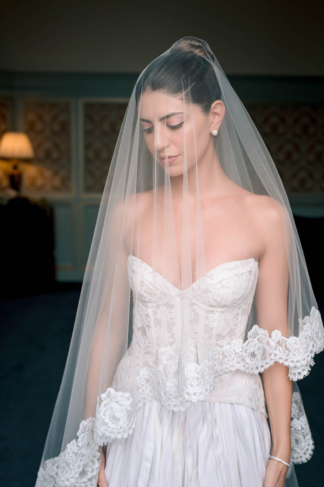 Bride in strapless lace corset gown and cathedral-length lace-trimmed veil looking down indoors