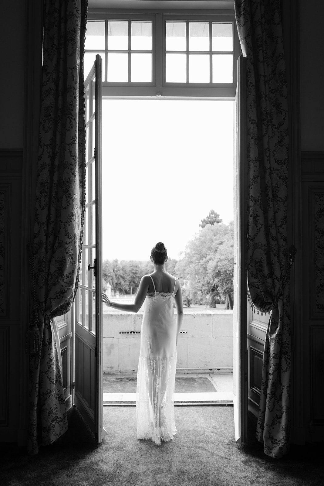 Black and white bride in spaghetti-strap lace gown standing at open French doors in chateau