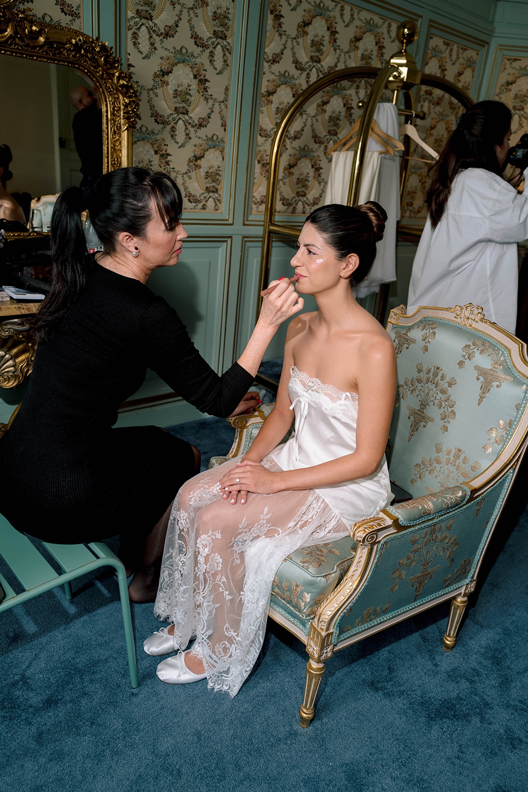 Makeup artist applying lipstick to bride in lace slip seated in gold-trimmed sage armchair