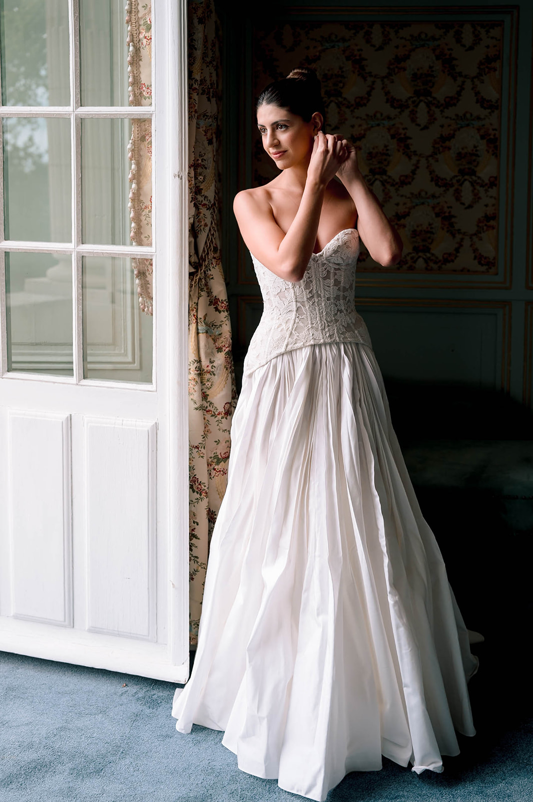 Bride adjusting earring in strapless lace corset gown in blue-grey paneled chateau room