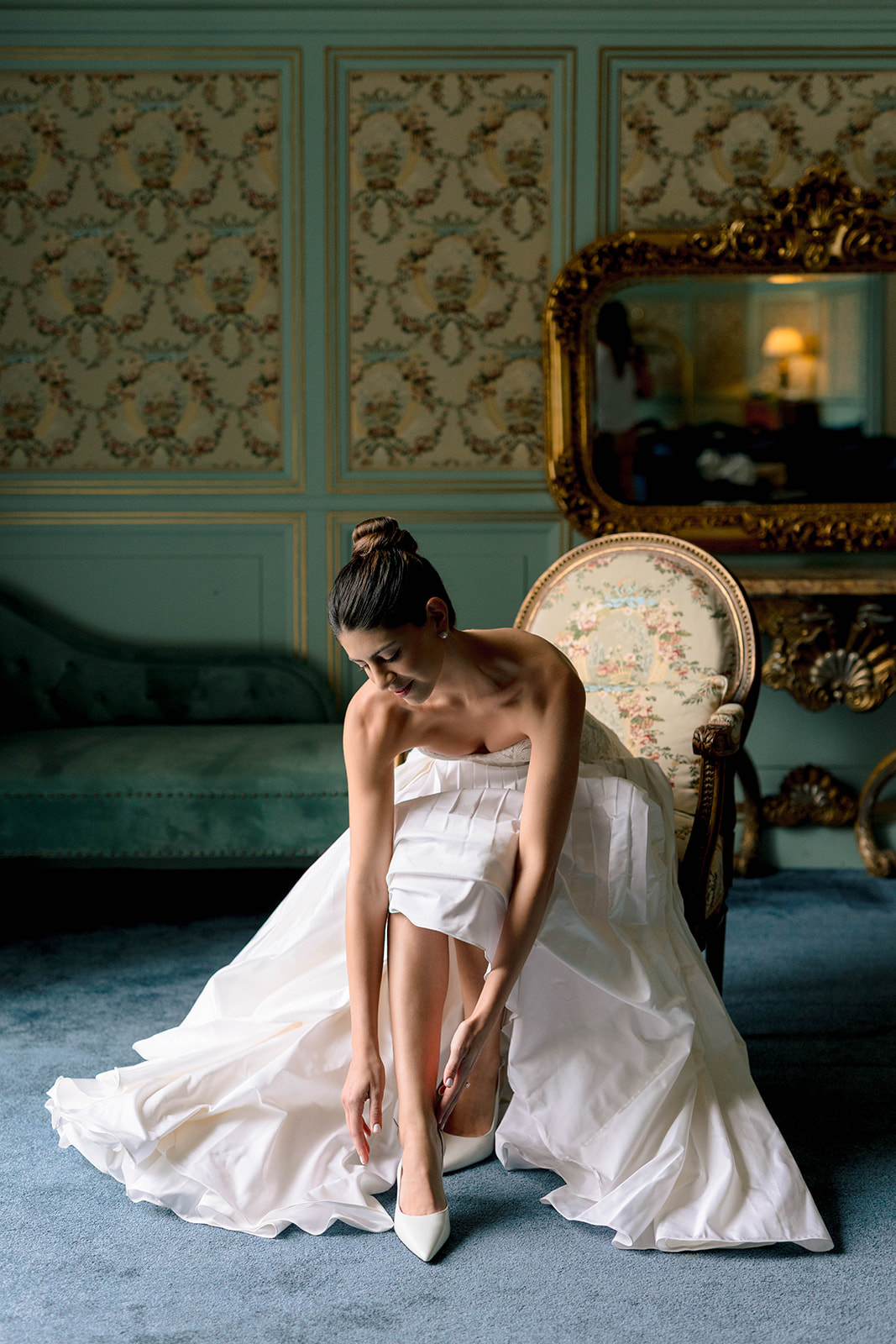 Bride fastening white pointed-toe heels in an ornate sage green and gold French chateau bridal suite
