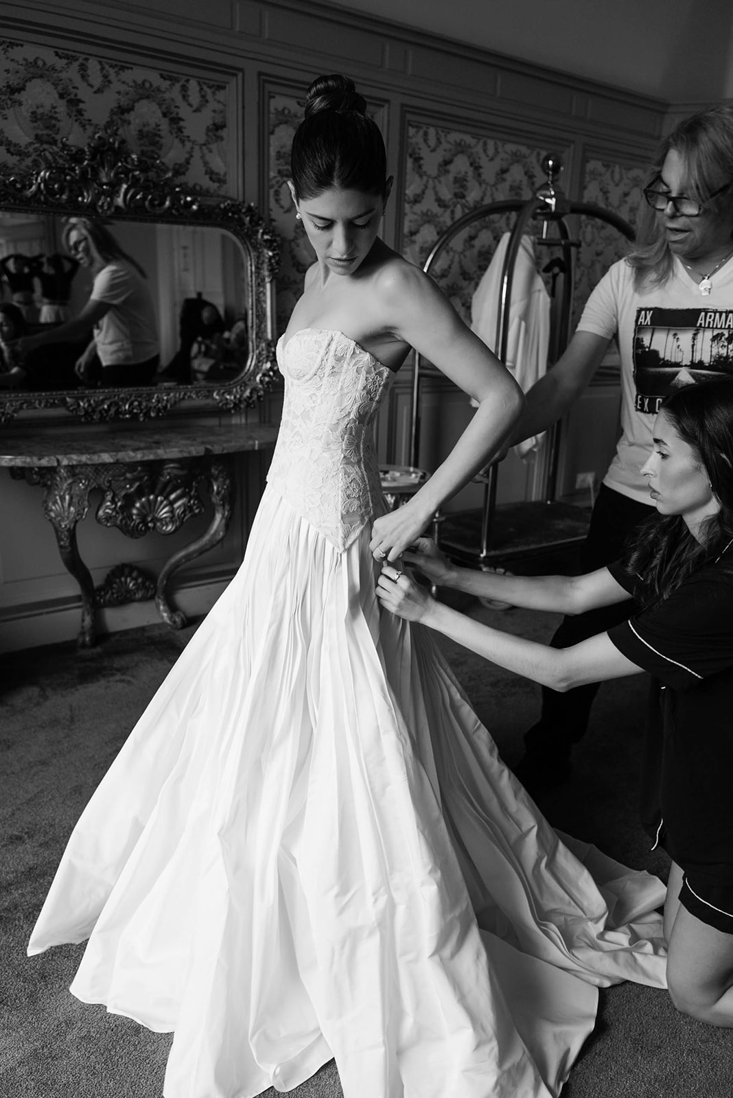 Black and white of bride in strapless lace ballgown being dressed by two attendants in gilt-furnished chateau room