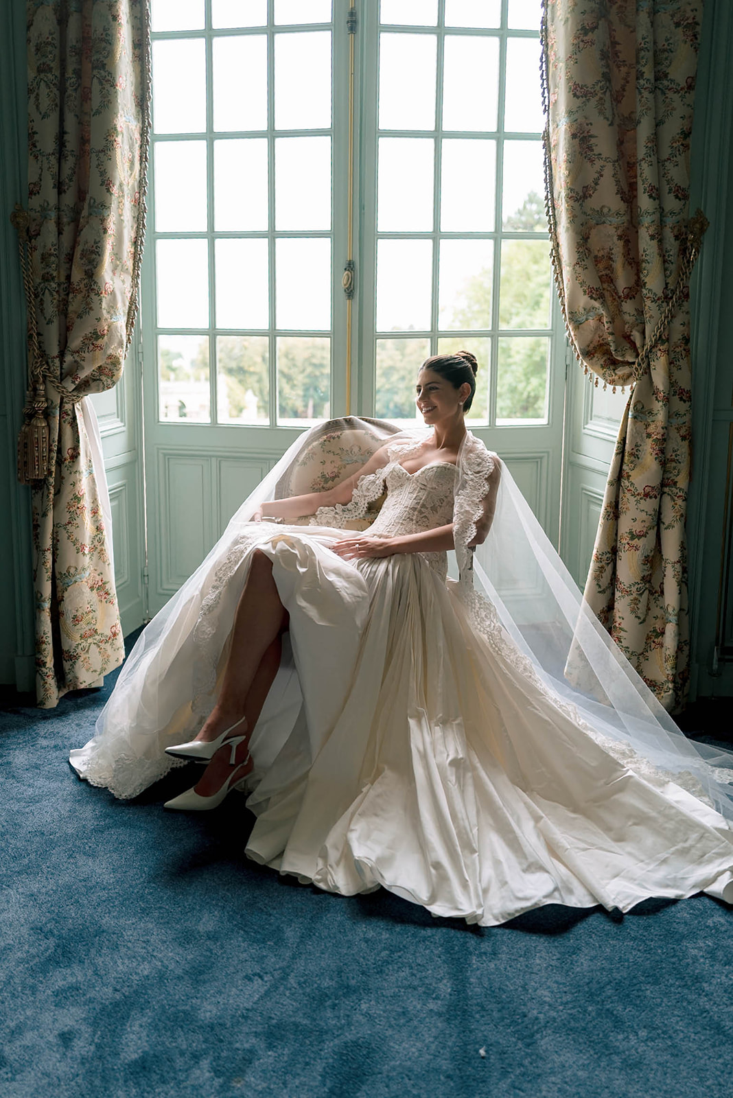 Bride seated in ballgown with cathedral lace veil spread across floor in chateau getting-ready room