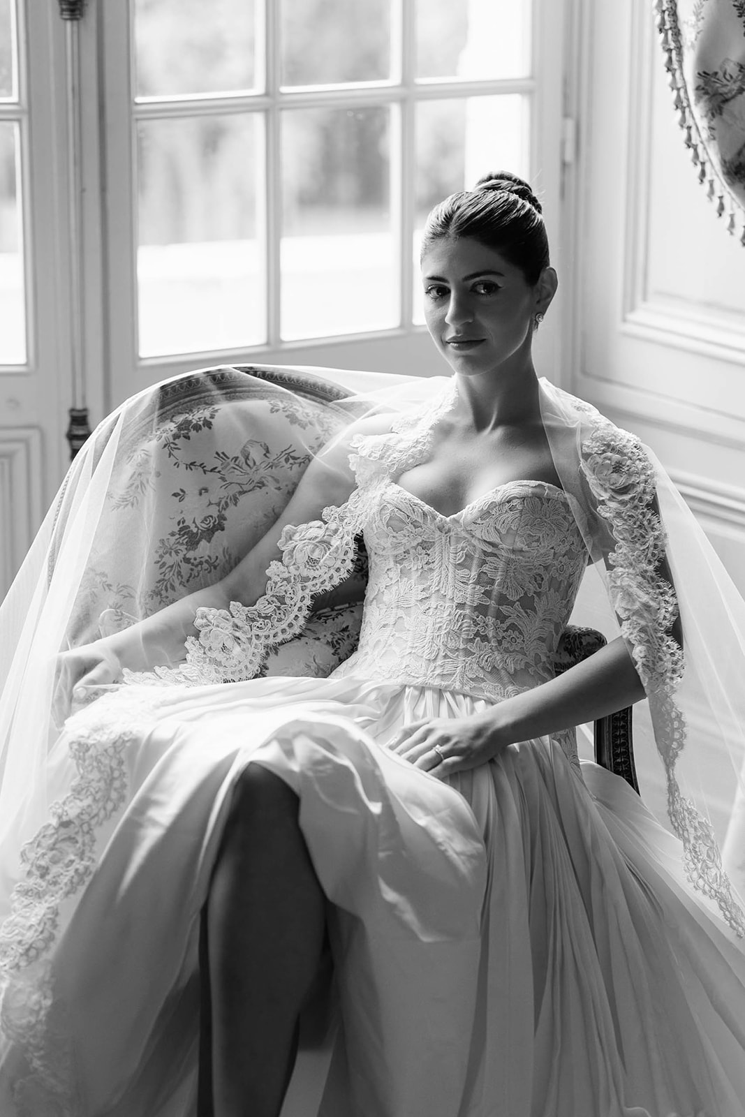 Black-and-white bridal portrait in ornate armchair with backlit French windows, bride in lace corset gown and long veil