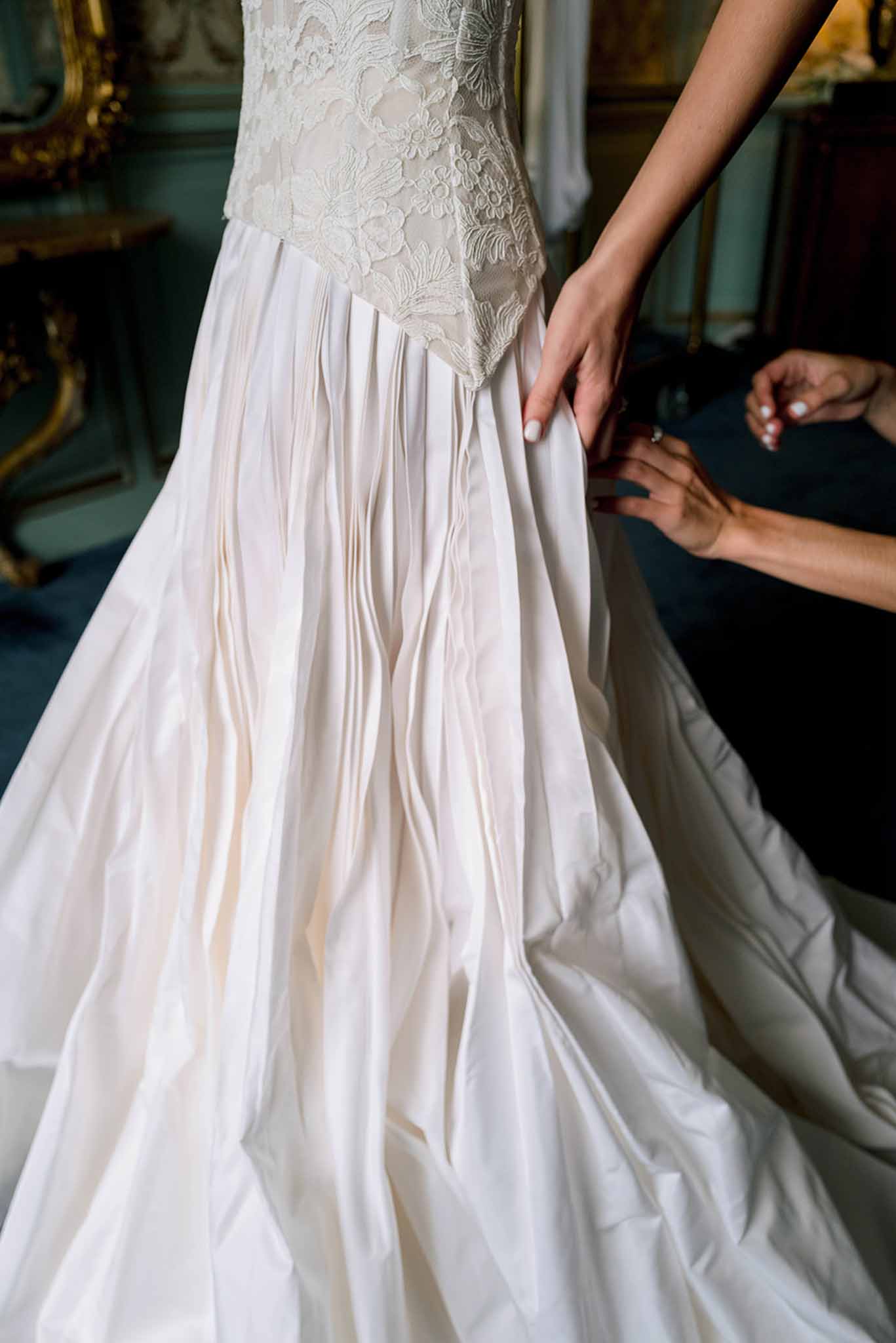 Hands fastening the back of a bride's ivory lace and satin wedding dress in a chateau dressing room